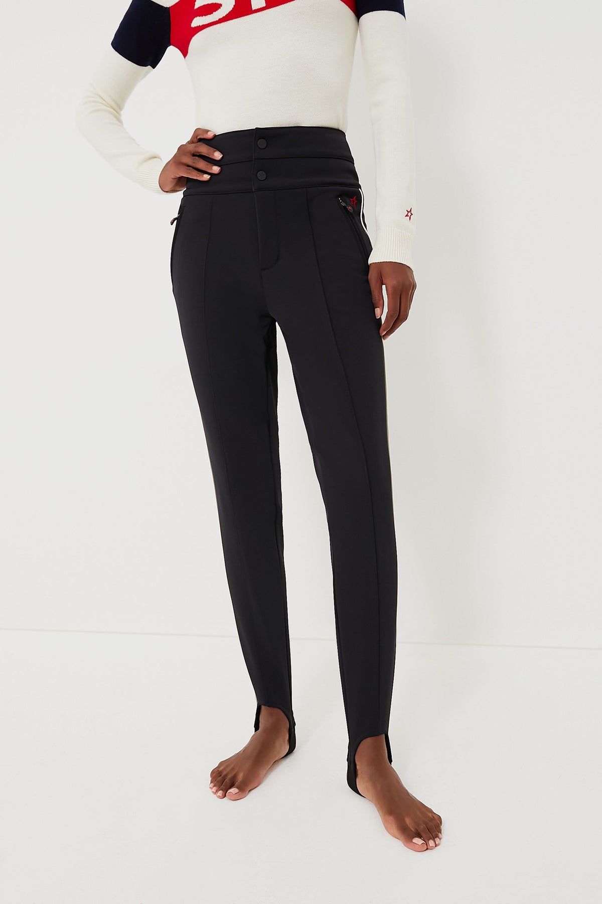 A person stands barefoot in Perfect Moment high-waisted black ski race pants with front buttons and a long-sleeve white, red, and navy top.