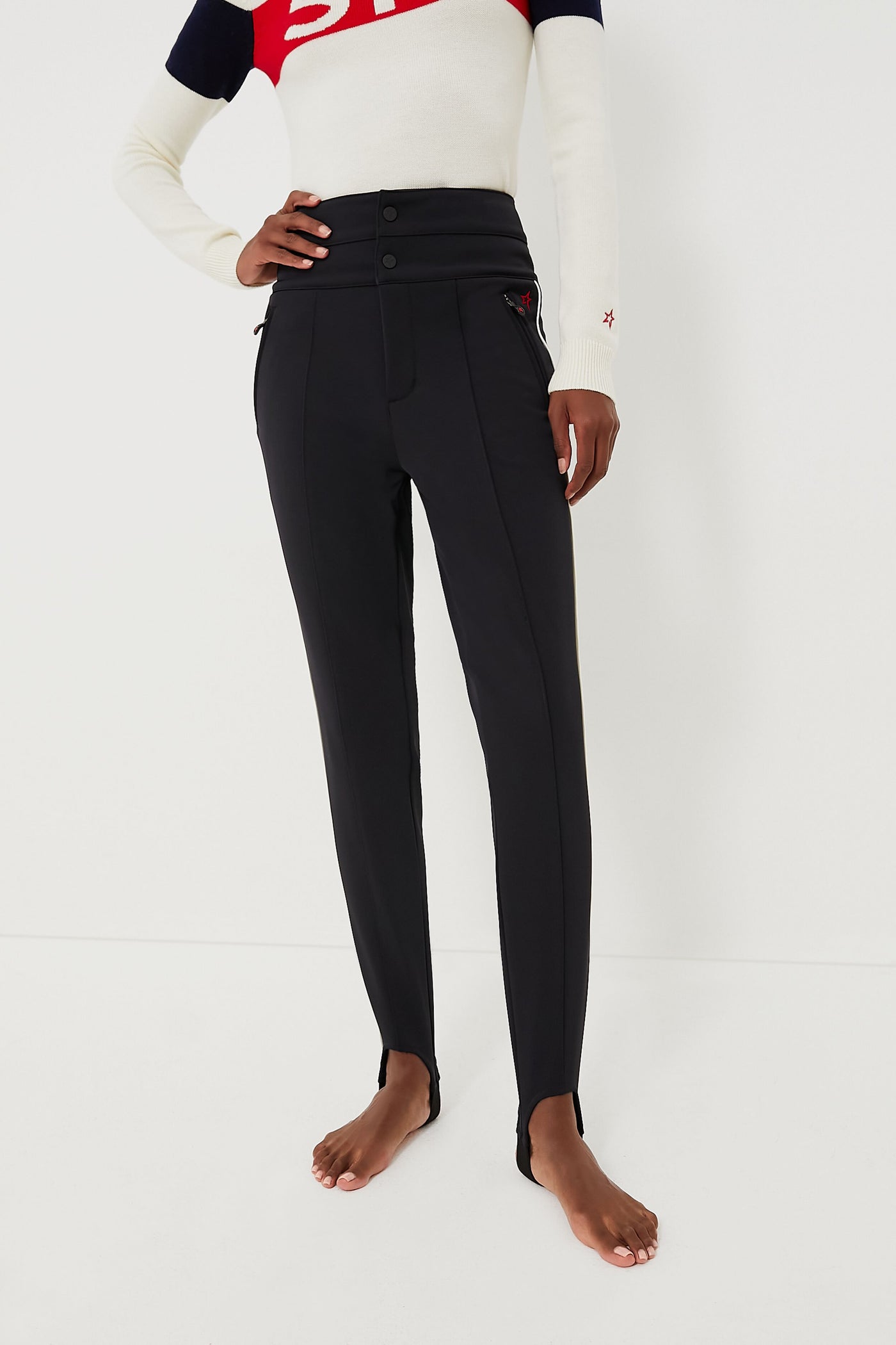A person stands barefoot in Perfect Moment high-waisted black ski race pants with front buttons and a long-sleeve white, red, and navy top.