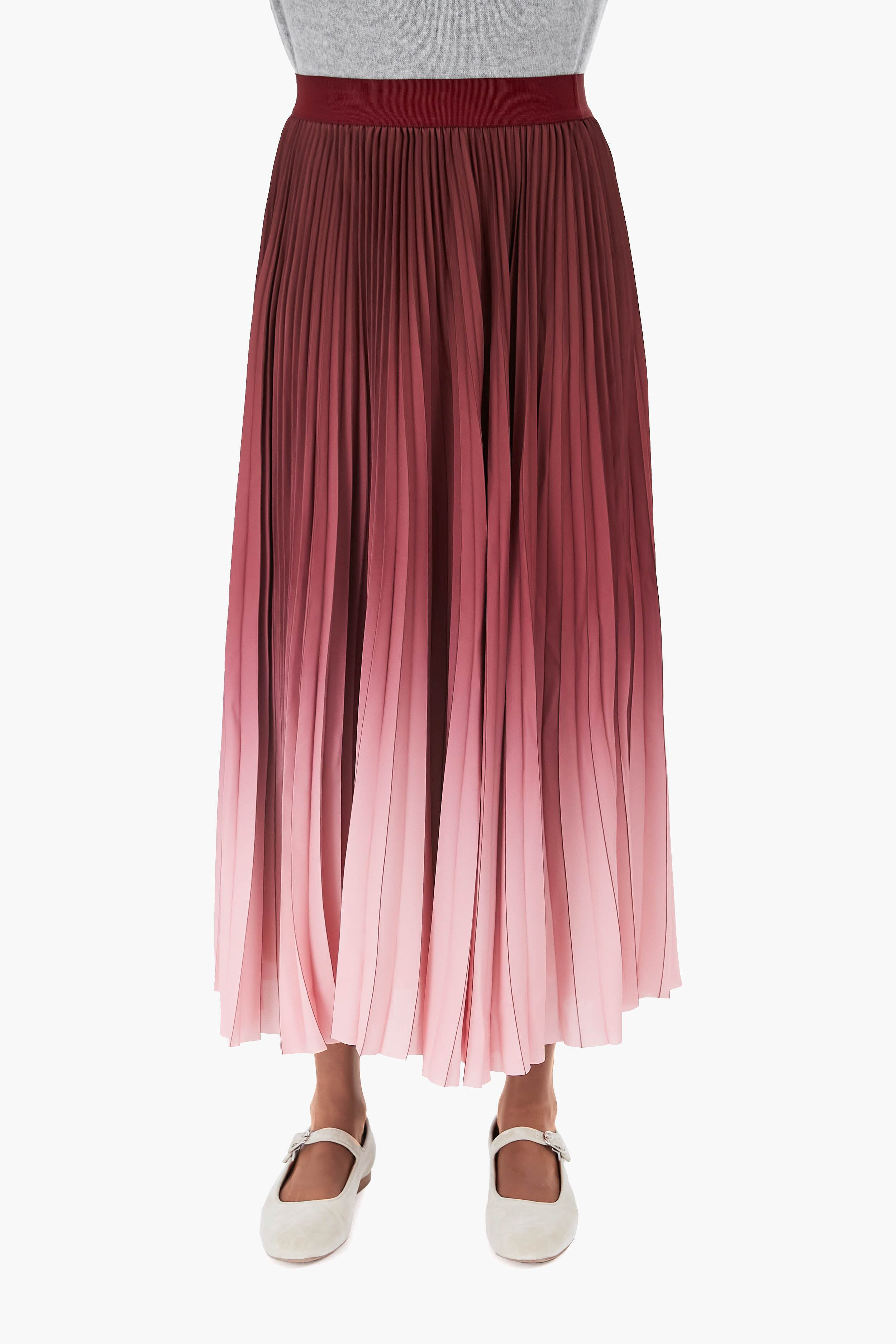 A person wears a gray top, a pleated midi skirt with a pink and burgundy gradient print by Weekend Max Mara, and white strapped shoes.