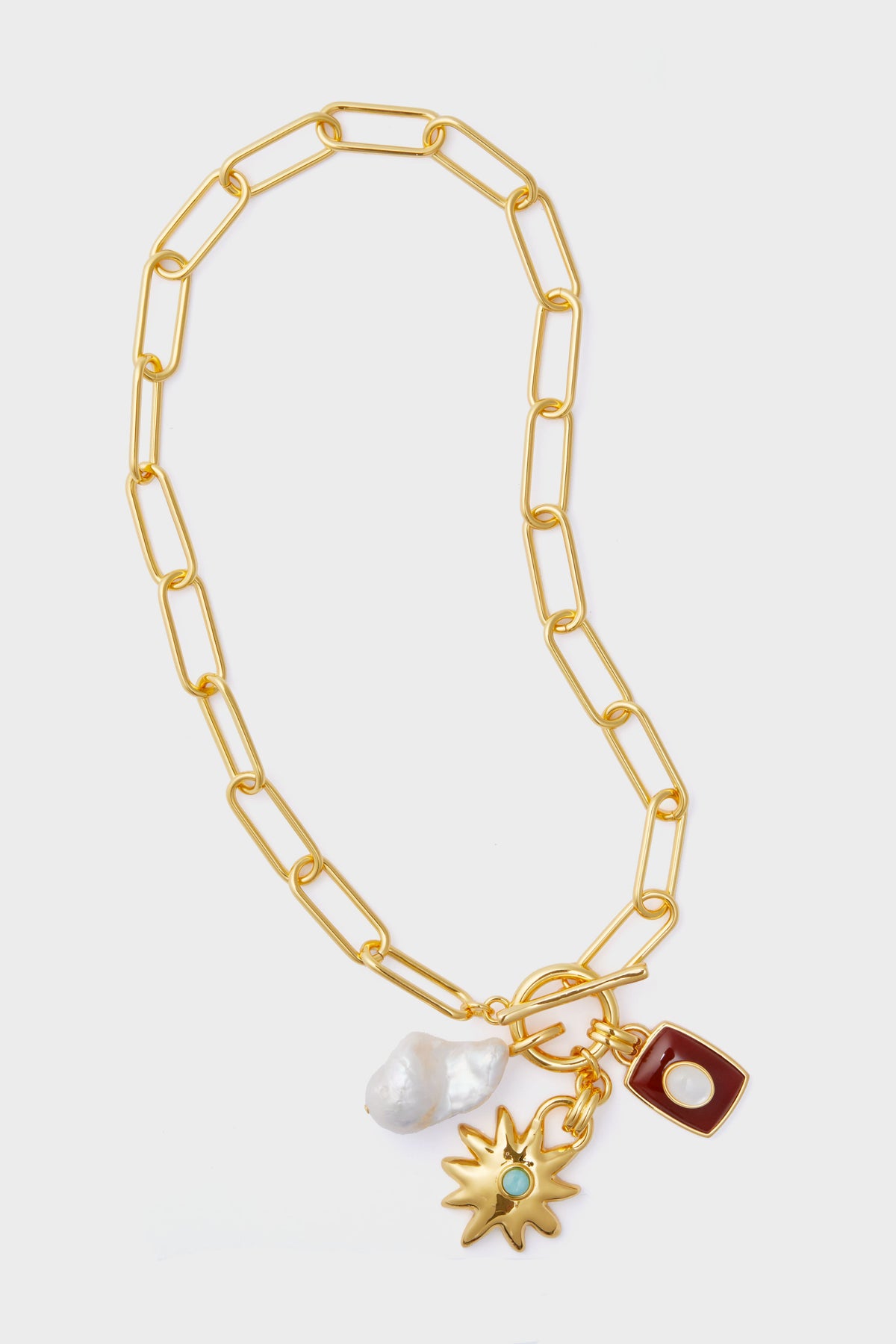 The Lizzie Fortunato Helios Charm Necklace is a handcrafted gold chain with large links, featuring three pendants: an irregular white pearl, a gold sunburst with a green gem, and a red rectangle with a pearl center.