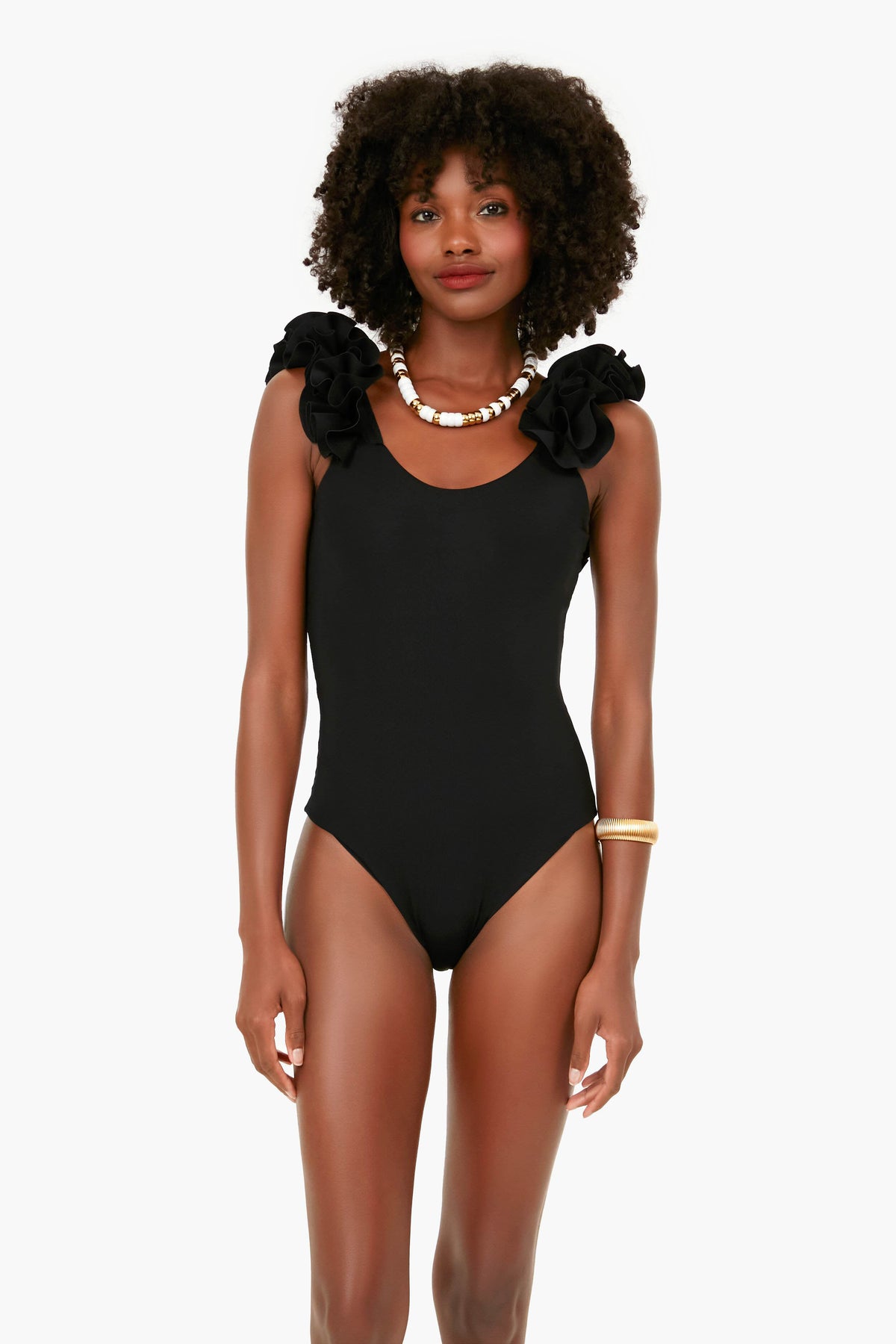 A woman wears a Maygel Coronel black one-piece swimsuit with ruffled shoulders, paired with a shell necklace and gold bangles.