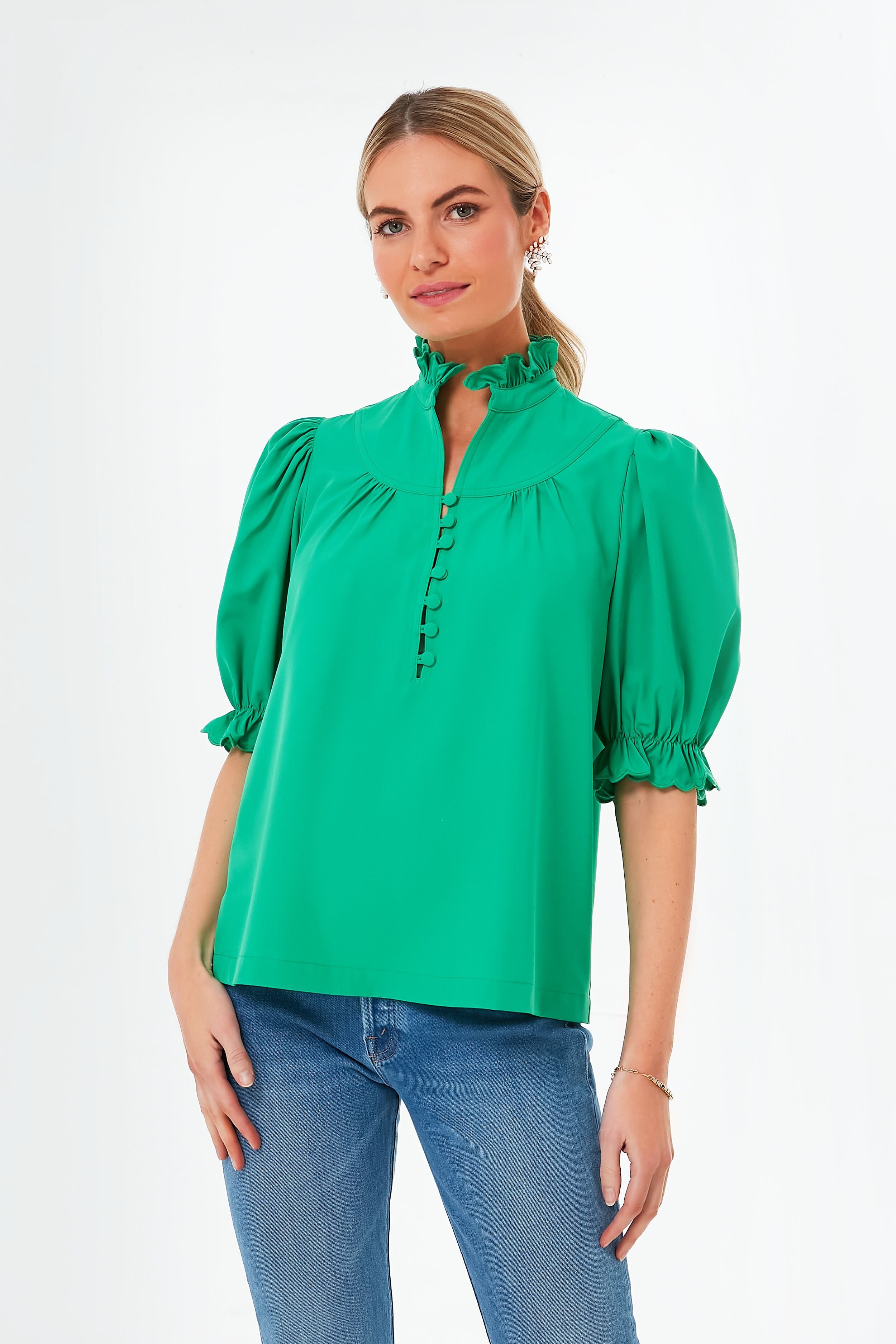 A woman wears a Pomander Place emerald green puff sleeve blouse with button details and a ruffle collar, paired with blue jeans.