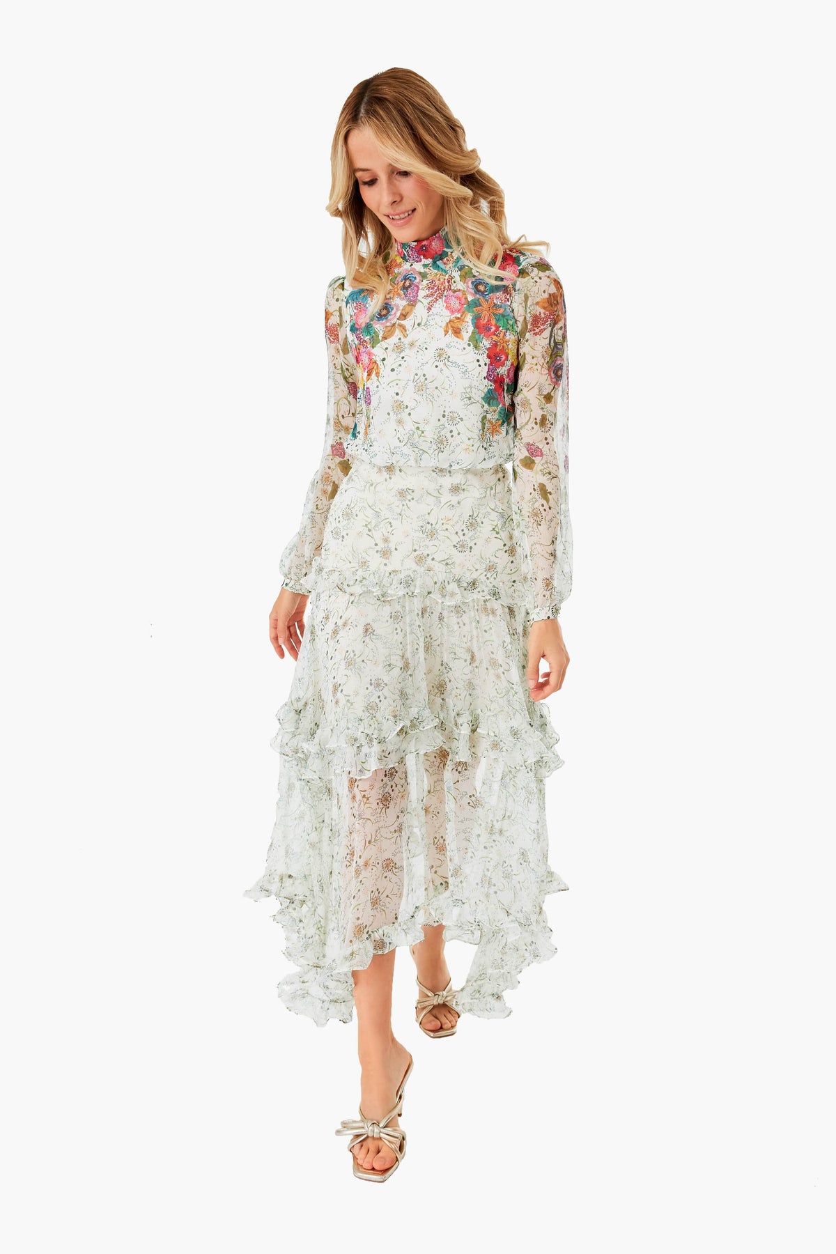 A woman wears a Saloni long-sleeved, high-neck garden party dress with a tiered ruffle skirt and open-toe heels, highlighting elegant floral style.