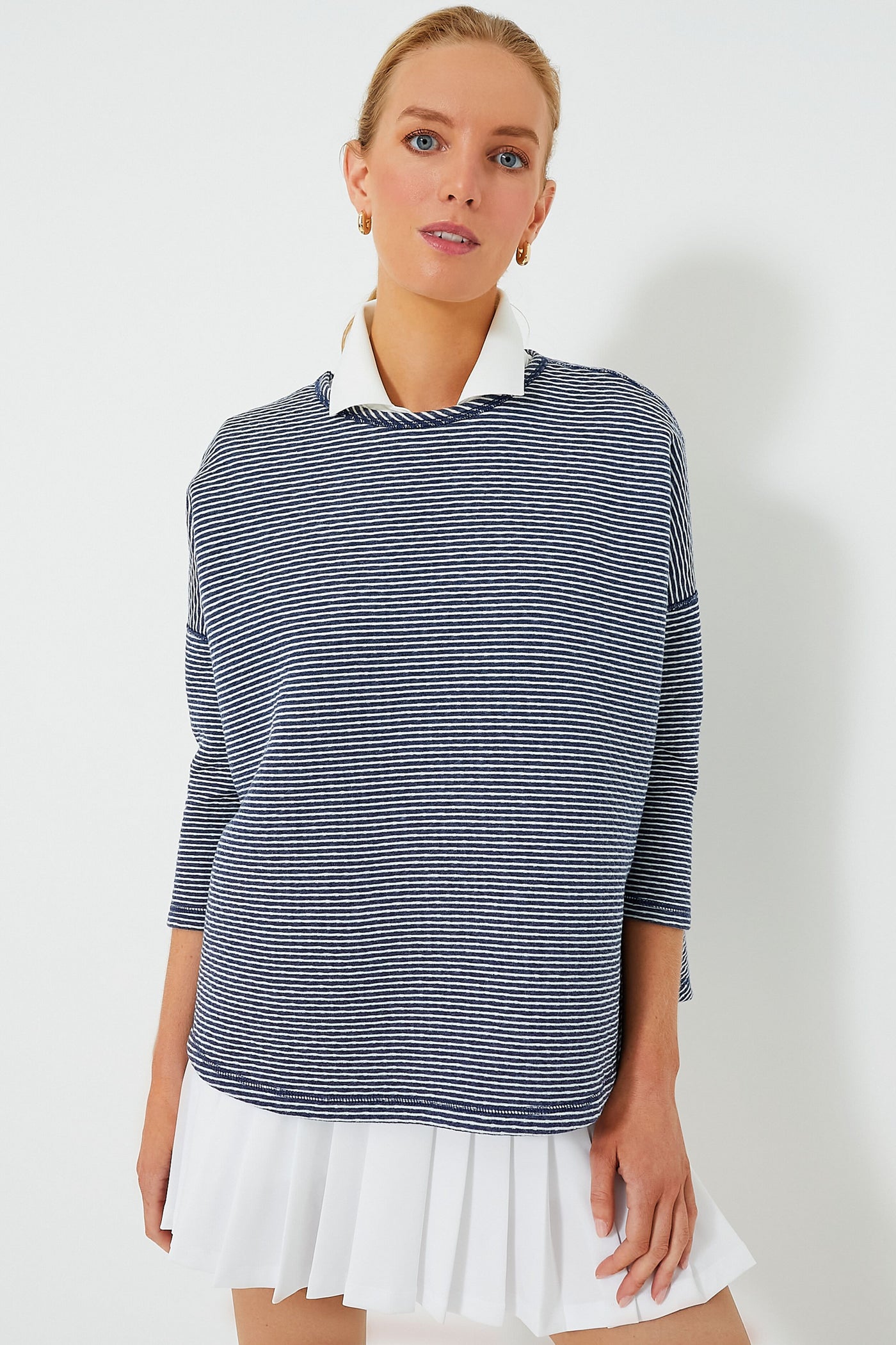 A woman wears a Tnuck Sport navy and white striped long-sleeve top layered over a white collared shirt and pleated skirt, featuring quilted fabric for a relaxed fit that combines comfort and style.