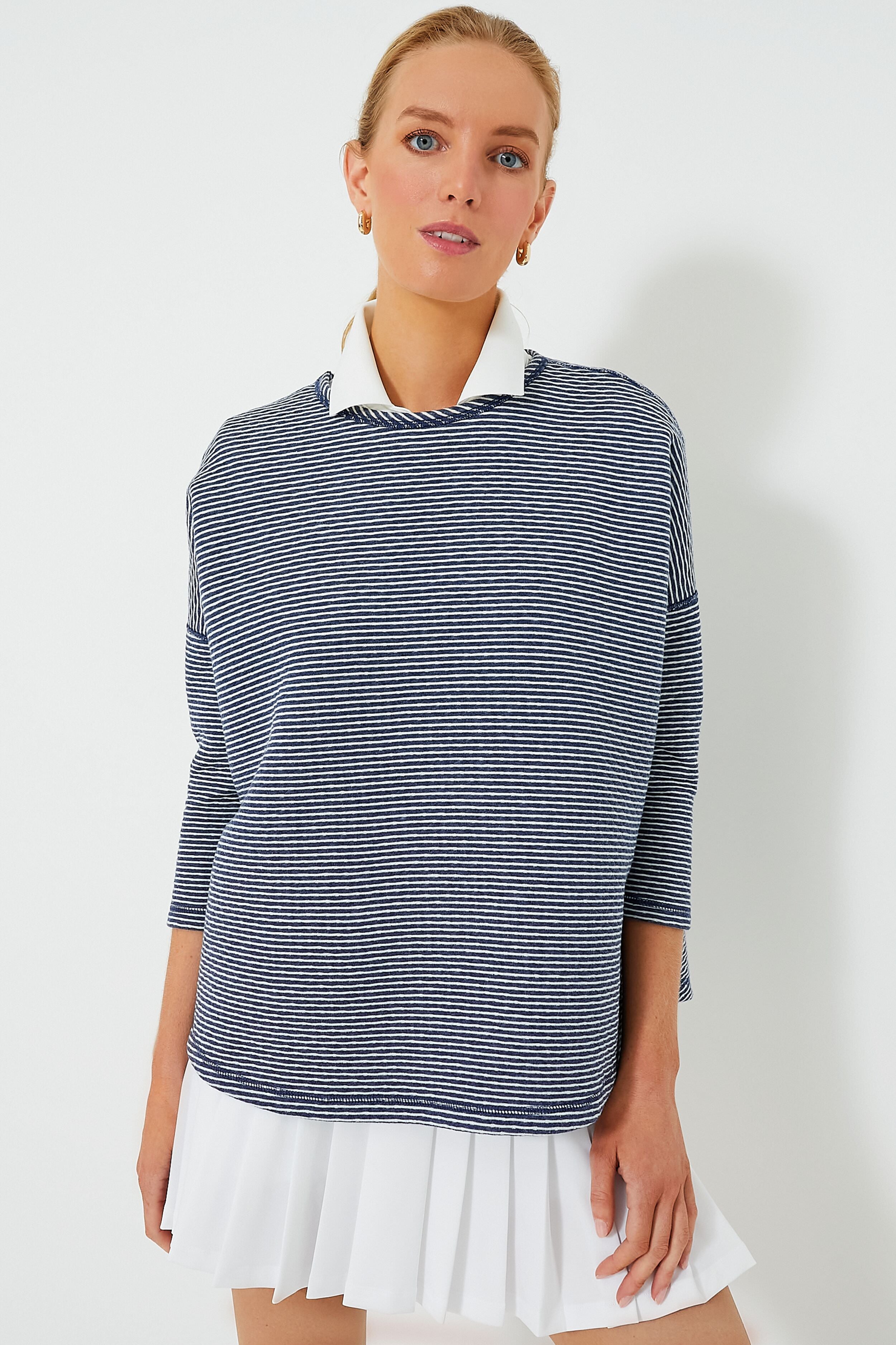 A woman wears a Tnuck Sport navy and white striped long-sleeve top layered over a white collared shirt and pleated skirt, featuring quilted fabric for a relaxed fit that combines comfort and style.