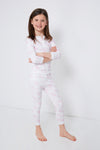A young girl stands barefoot against a white wall, smiling with her arms crossed in Nella Pima Pink Toile Baby Pajamas, crafted from the softest Pima cotton with light pink and white long sleeves.