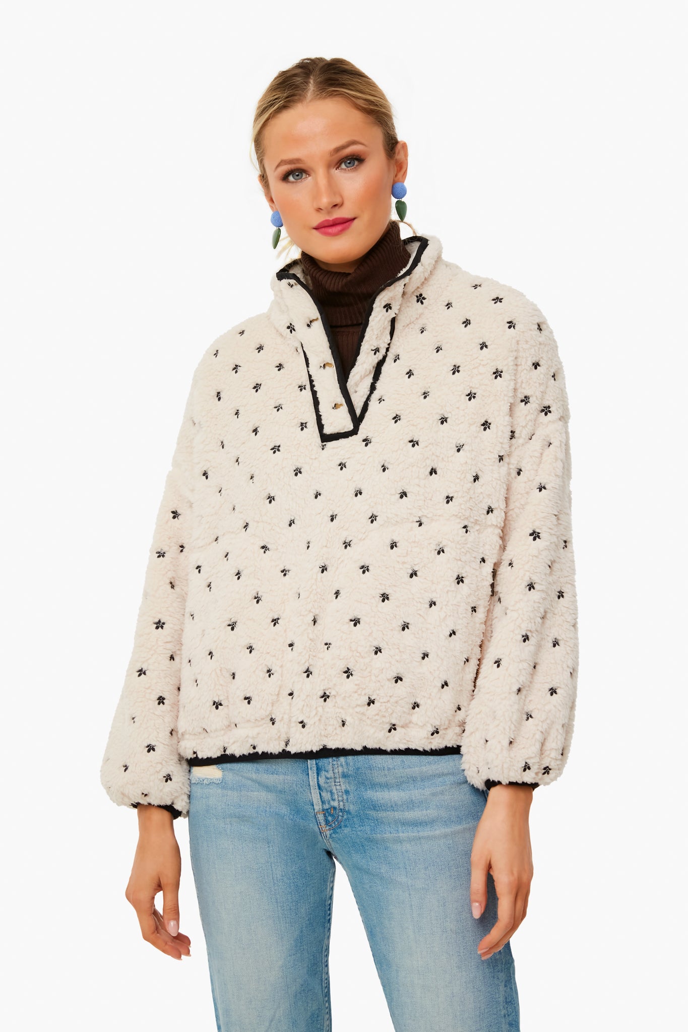Woman wearing an embroidered fleece jacket with black star patterns by THE GREAT., a brown turtleneck, blue jeans, and colorful earrings.