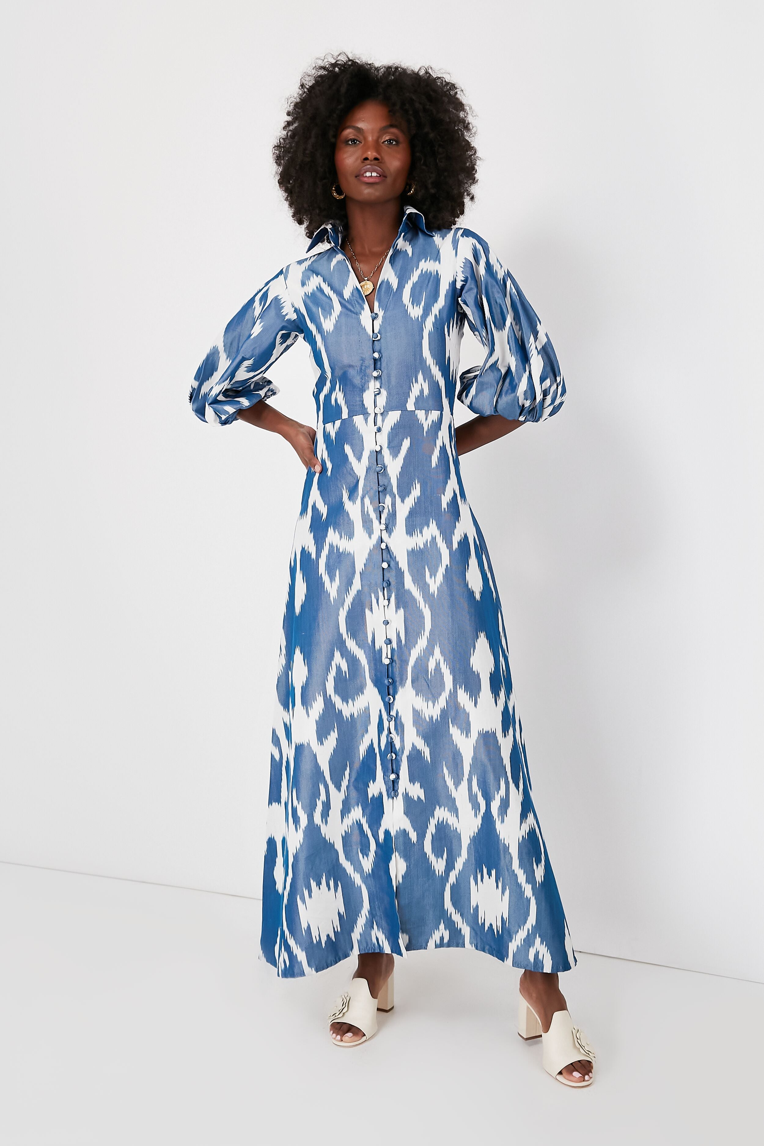 A woman wears a blue and white patterned silk maxi dress with puffed sleeves and handmade embroidery by De Castro, paired with white open-toe heeled sandals.