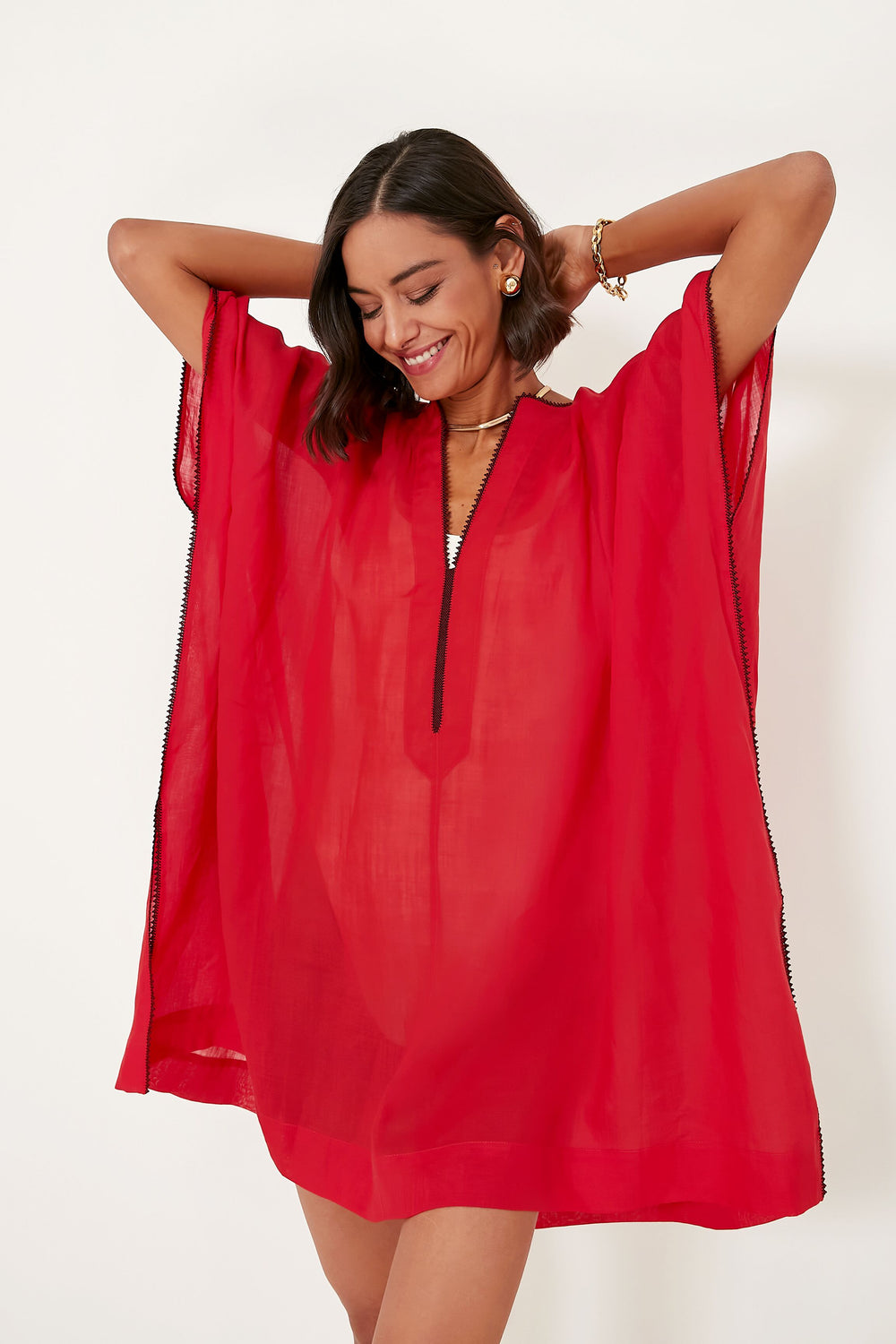 A woman smiles and raises her arms in a loose, sheer red dress with short sleeves by Three Graces London—perfect as a beach kaftan or oversized coverup for sustainable resort wear.