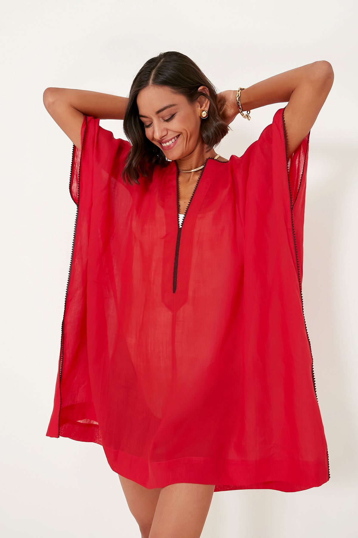 A woman smiles and raises her arms in a loose, sheer red dress with short sleeves by Three Graces London—perfect as a beach kaftan or oversized coverup for sustainable resort wear.