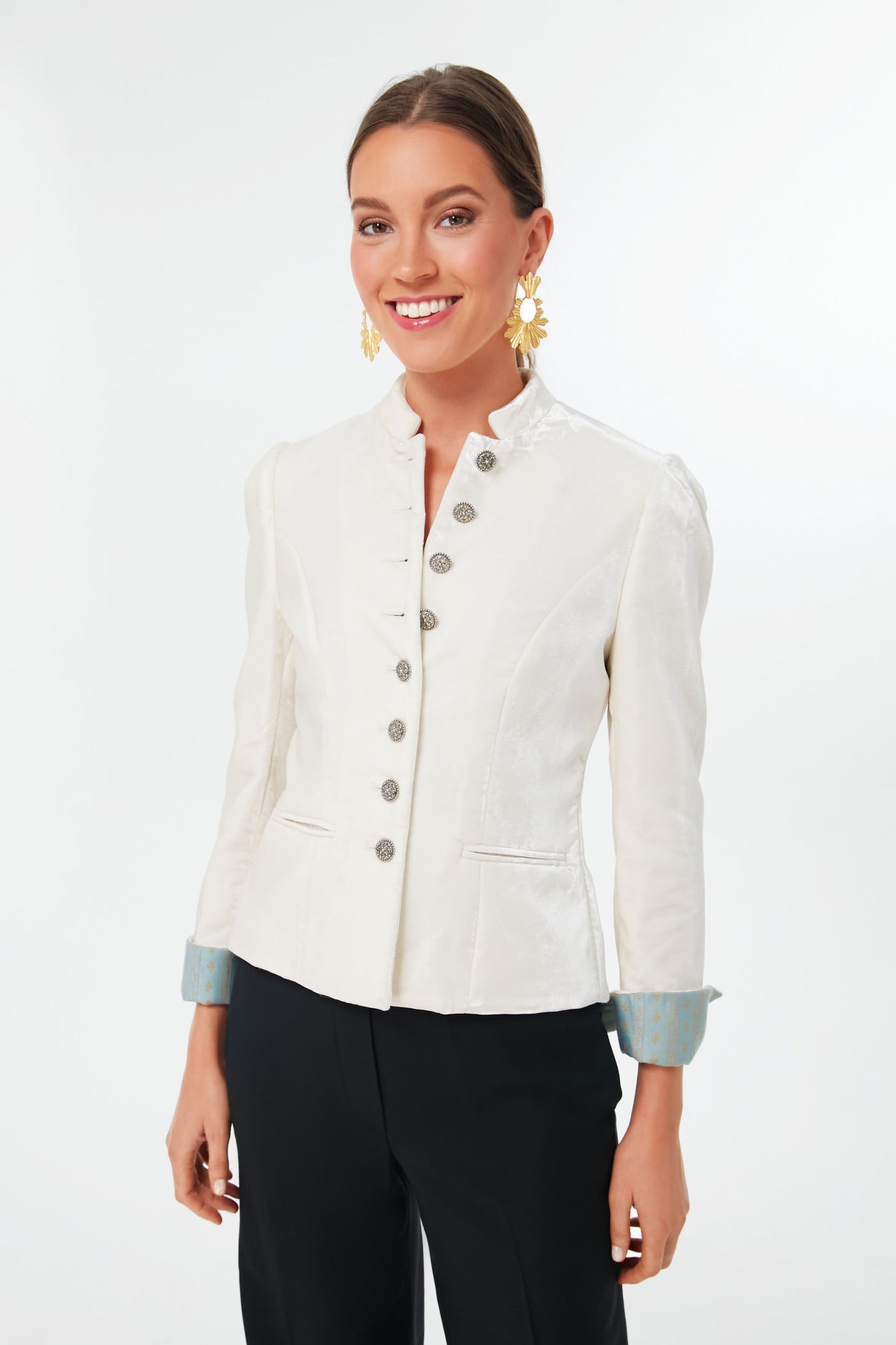 A woman confidently smiles in a vintage-inspired white buttoned jacket with light blue cuffs from Sanoe Collection, paired with black pants and gold statement earrings.