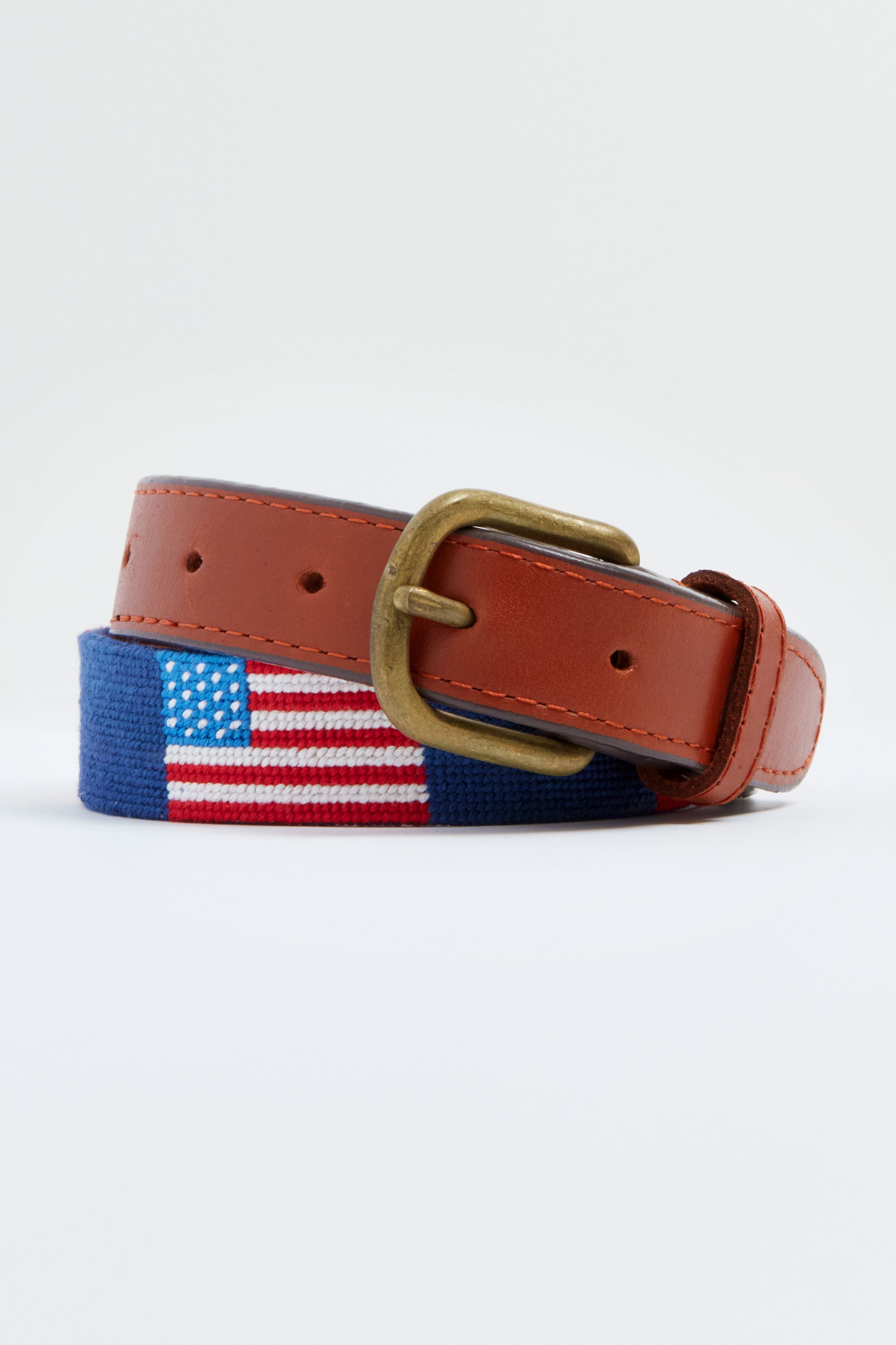 A Smathers and Branson kids belt made of brown leather and blue fabric, hand-stitched with an embroidered American flag and finished with a brass buckle, is coiled neatly.