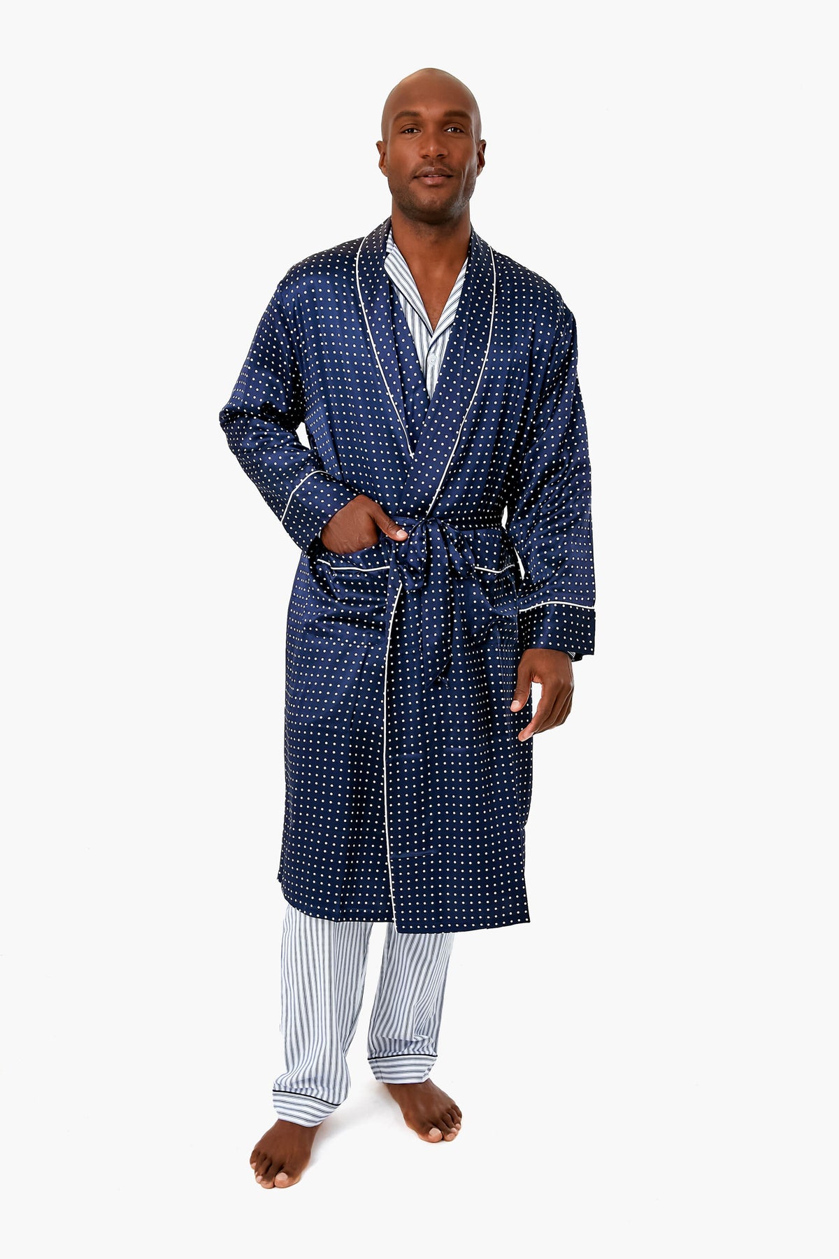A man stands barefoot in Petite Plume’s blue patterned luxury robe over striped pajamas, showcasing comfortable loungewear.
