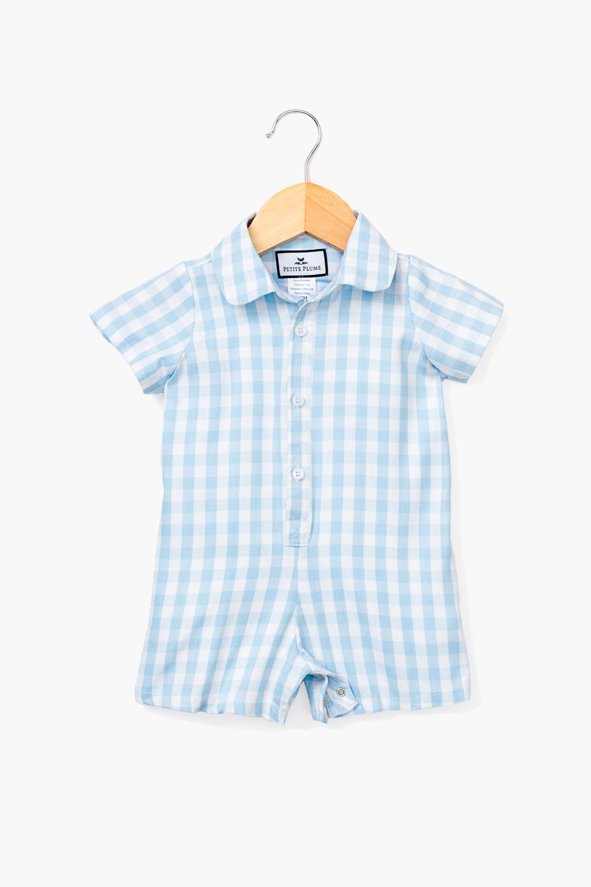 A Petite Plume short-sleeve blue gingham infant romper with a button front and collar, crafted from soft cotton sleepwear, is displayed on a wooden hanger.