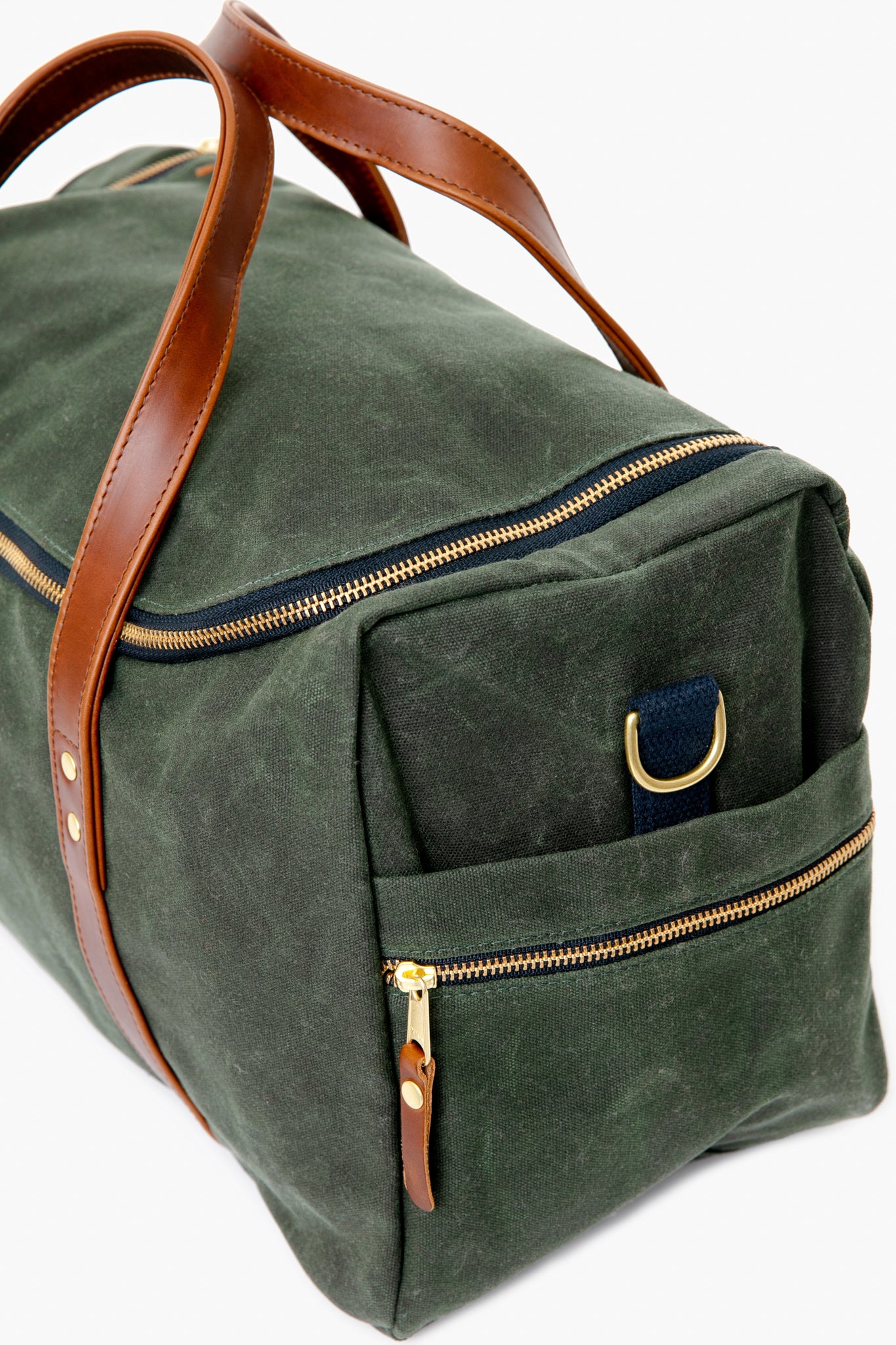 The Hudson Sutler green canvas commuter duffel features brown leather handles, gold zippers, and an exterior zip pocket. Made in the USA—perfect for fans of customizable bags.
