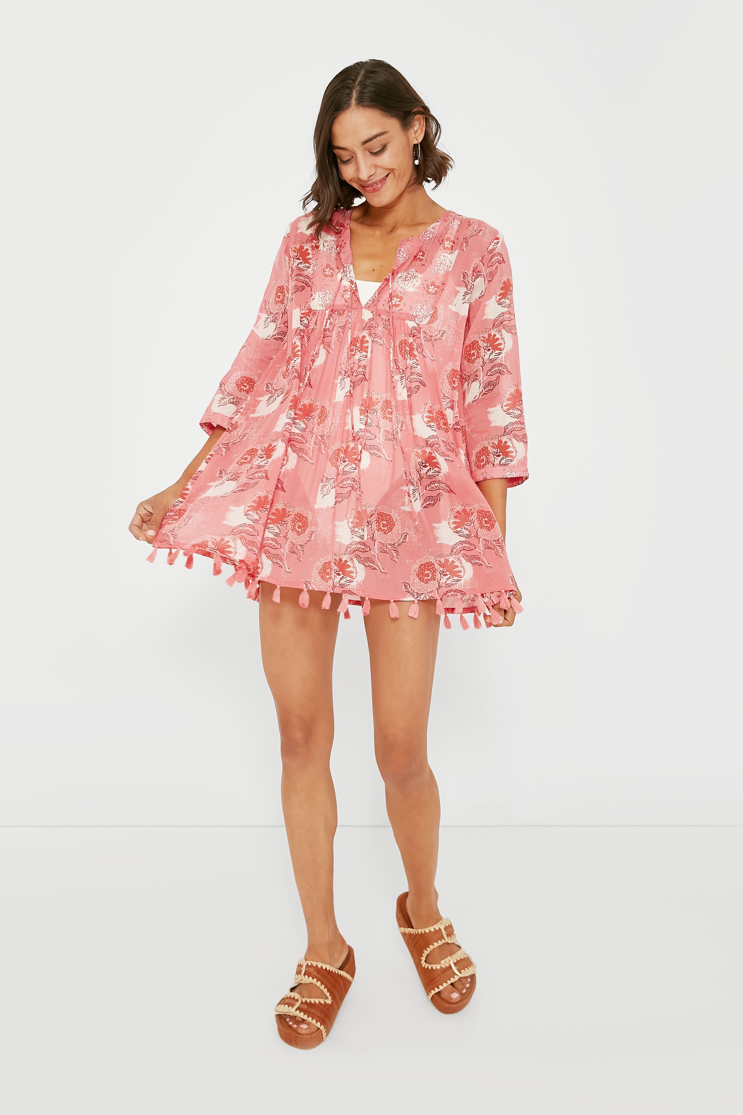 A woman in a pink floral tassel tunic dress by Ro's Garden, styled with brown sandals, holds the sides of her boho-chic cover-up—an ideal vacation-ready look for your next getaway.