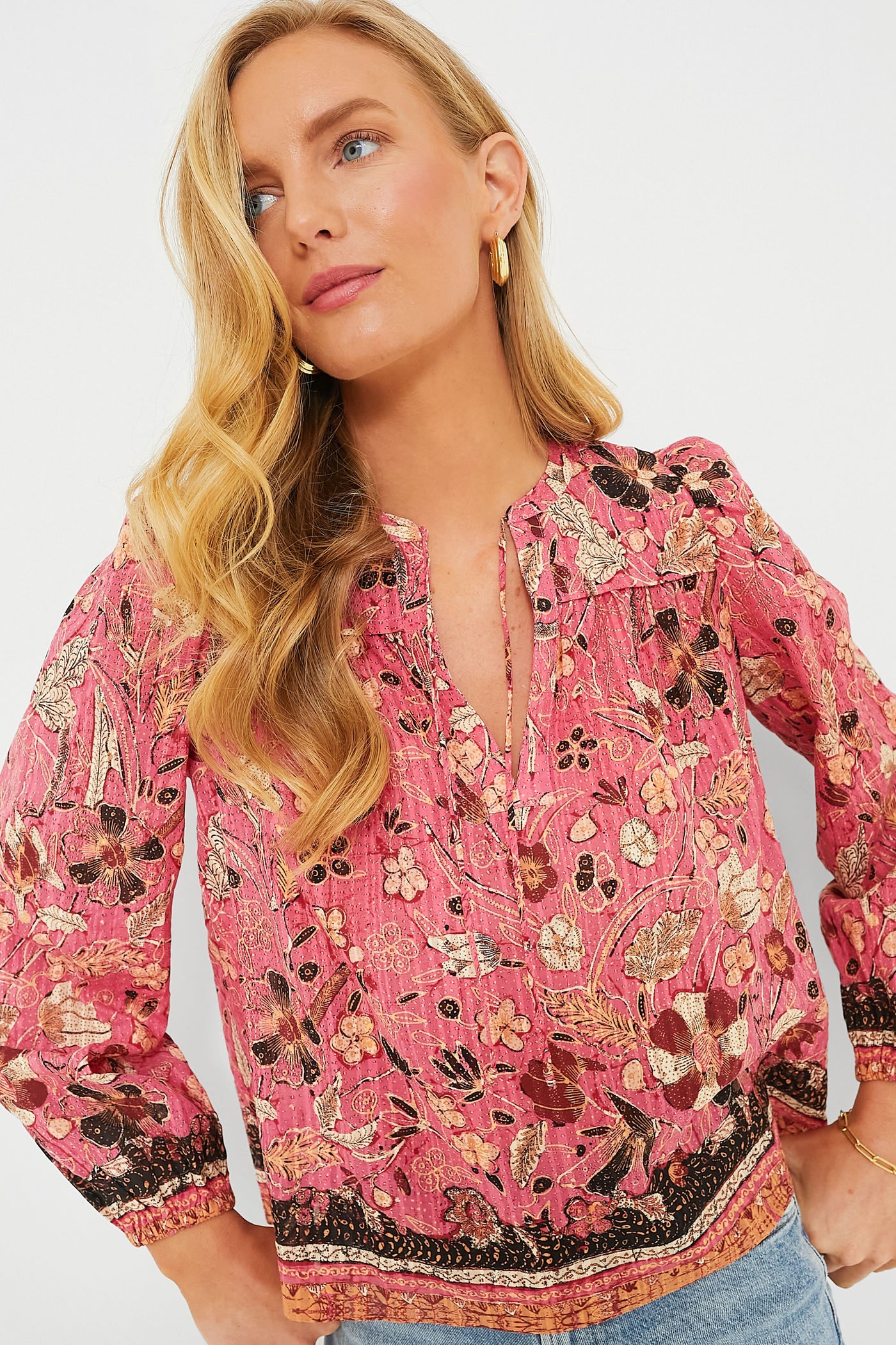 A woman wearing a pink floral Ulla Johnson blouse with blouson sleeves and a split neckline, styled with gold hoop earrings, poses with one hand on her hip.