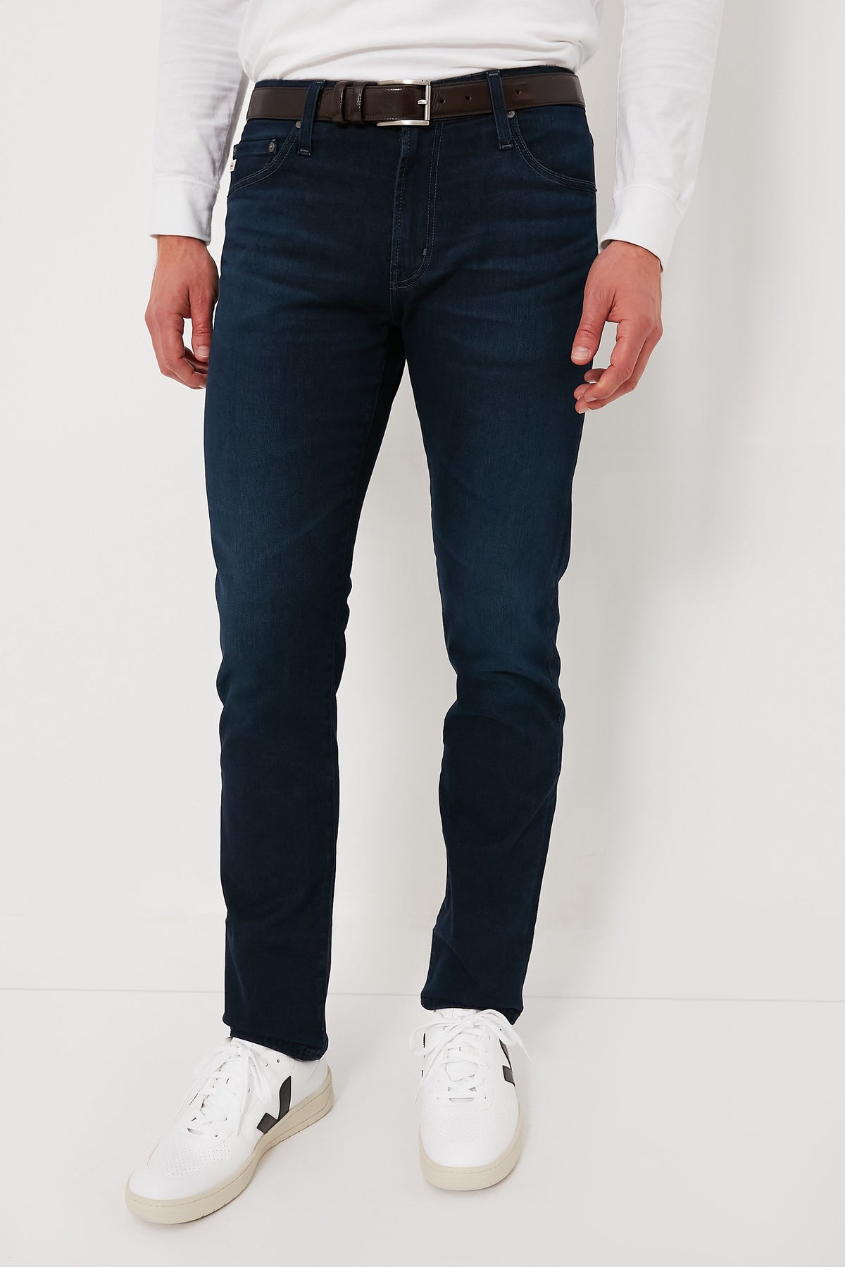 A man wears AG Jeans in dark blue premium denim, paired with a white long-sleeve shirt, white sneakers, and a brown belt.
