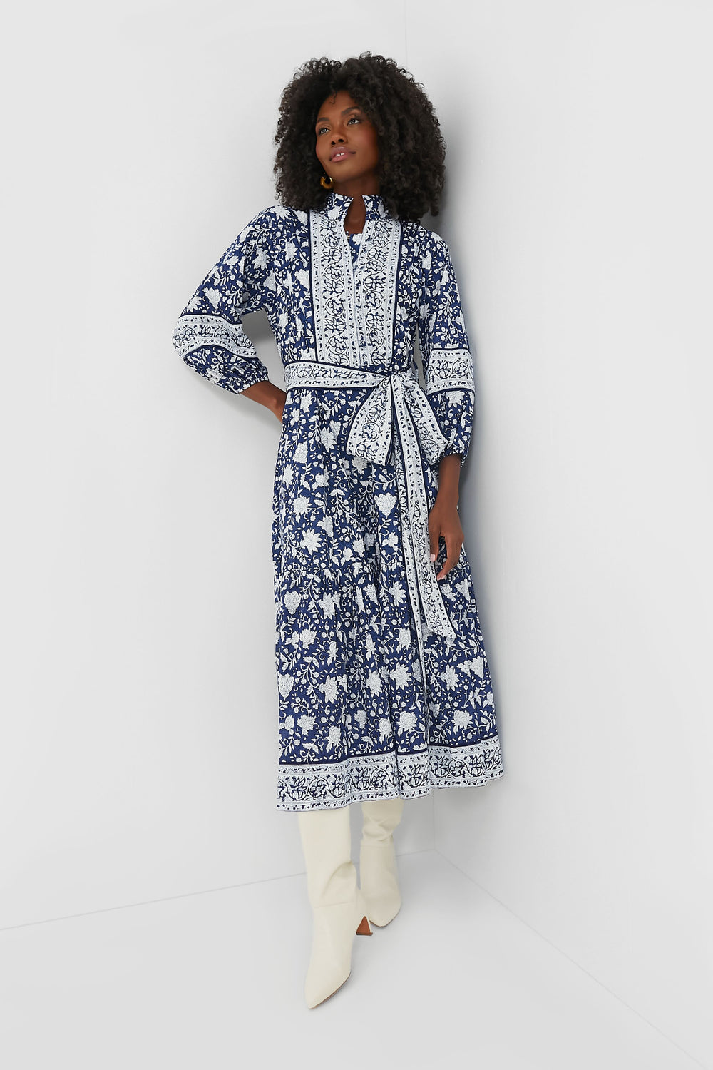 A woman wears the Sue Sartor Navy Flounce Dress, featuring blue and white block-printed florals and a belt, paired with white knee-high boots—crafted from sustainable fabrics.