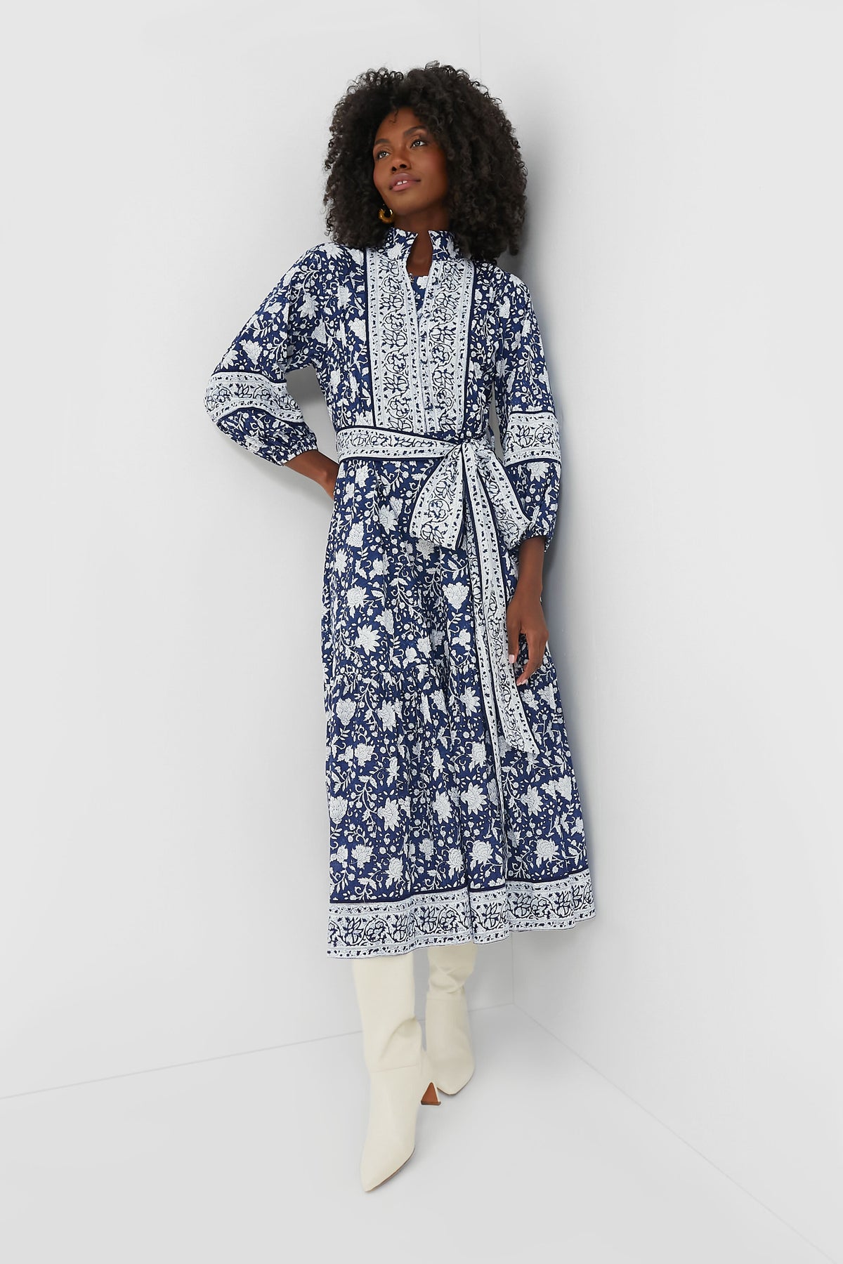 A woman wears the Sue Sartor Navy Flounce Dress, featuring blue and white block-printed florals and a belt, paired with white knee-high boots—crafted from sustainable fabrics.