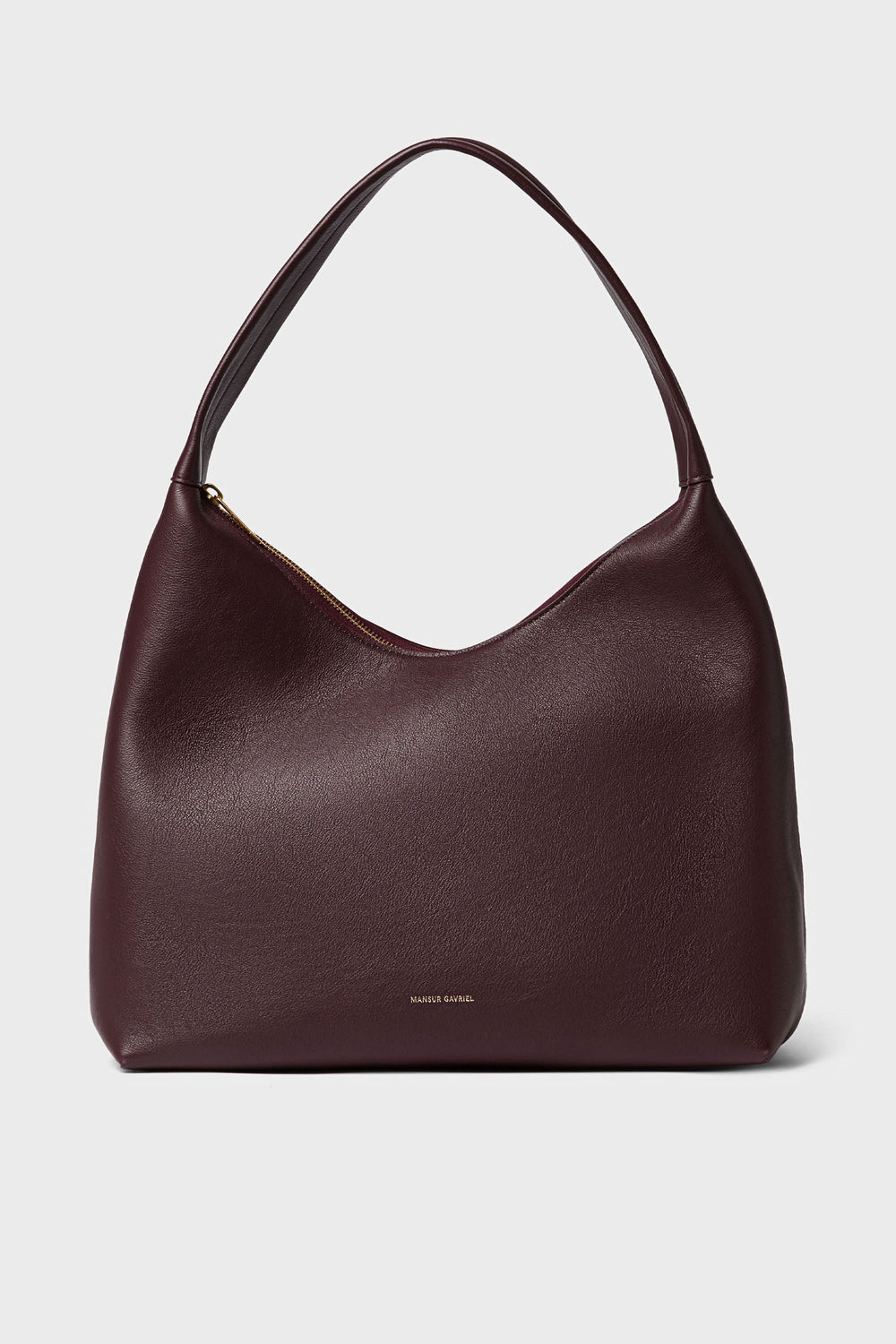 A Mansur Gavriel shoulder bag in Italian leather, burgundy color, showcases a curved top, wide single strap, subtle gold zipper closure, and small gold brand text near the bottom center.