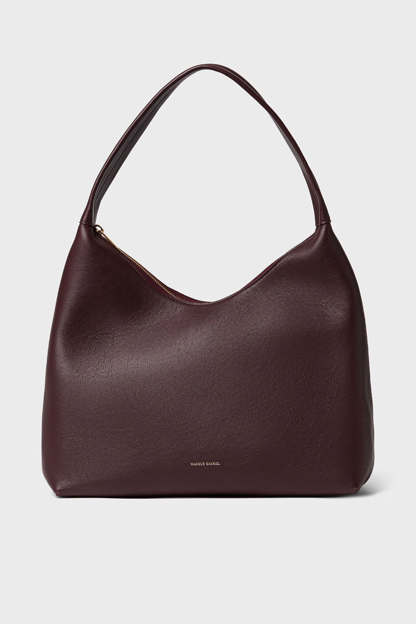 A Mansur Gavriel shoulder bag in Italian leather, burgundy color, showcases a curved top, wide single strap, subtle gold zipper closure, and small gold brand text near the bottom center.