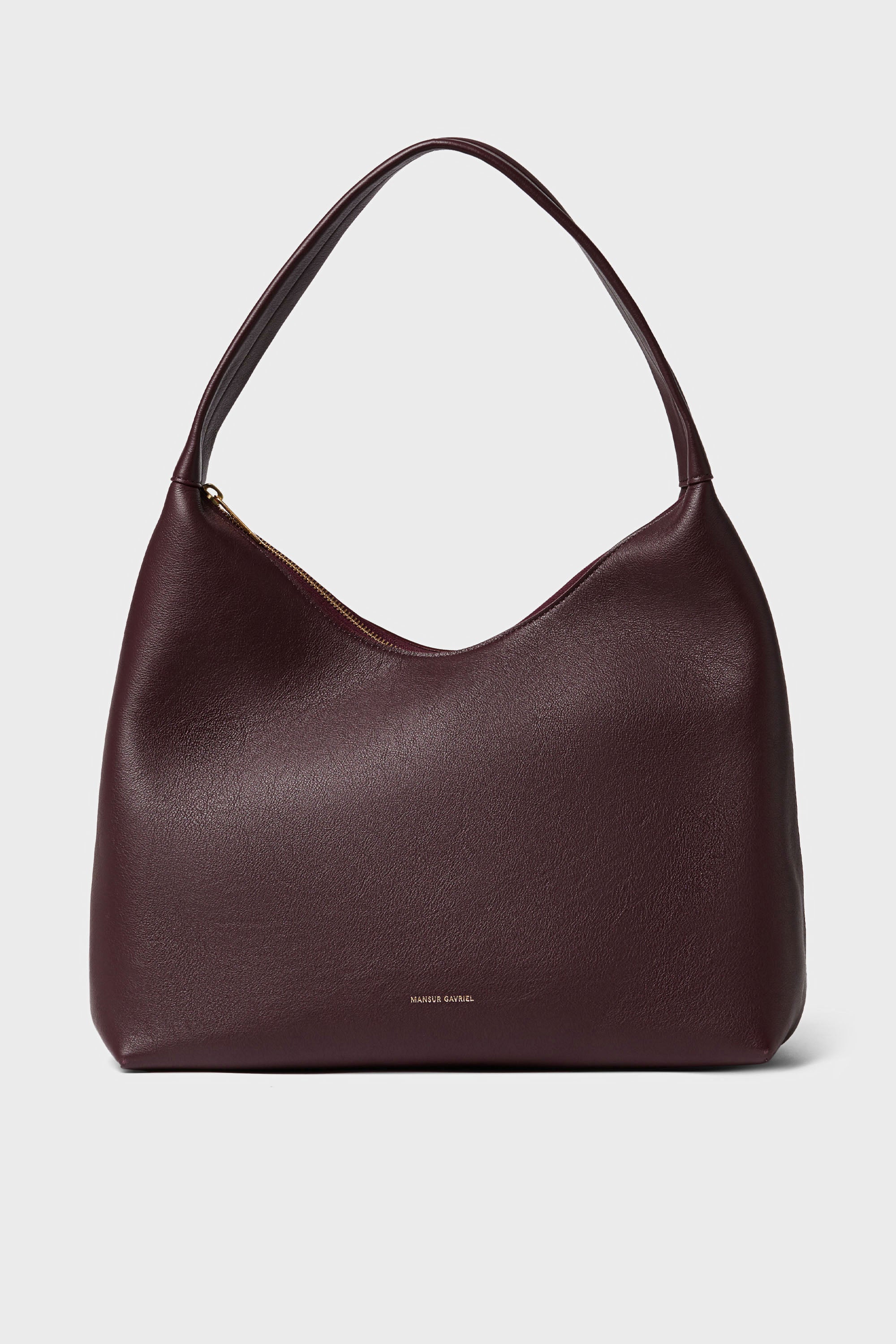 A Mansur Gavriel shoulder bag in Italian leather, burgundy color, showcases a curved top, wide single strap, subtle gold zipper closure, and small gold brand text near the bottom center.