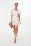 A woman wears a Hyacinth House light pink floral mini dress with puff sleeves, paired with a woven clutch and beige sandals.