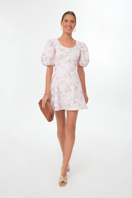 A woman wears a Hyacinth House light pink floral mini dress with puff sleeves, paired with a woven clutch and beige sandals.