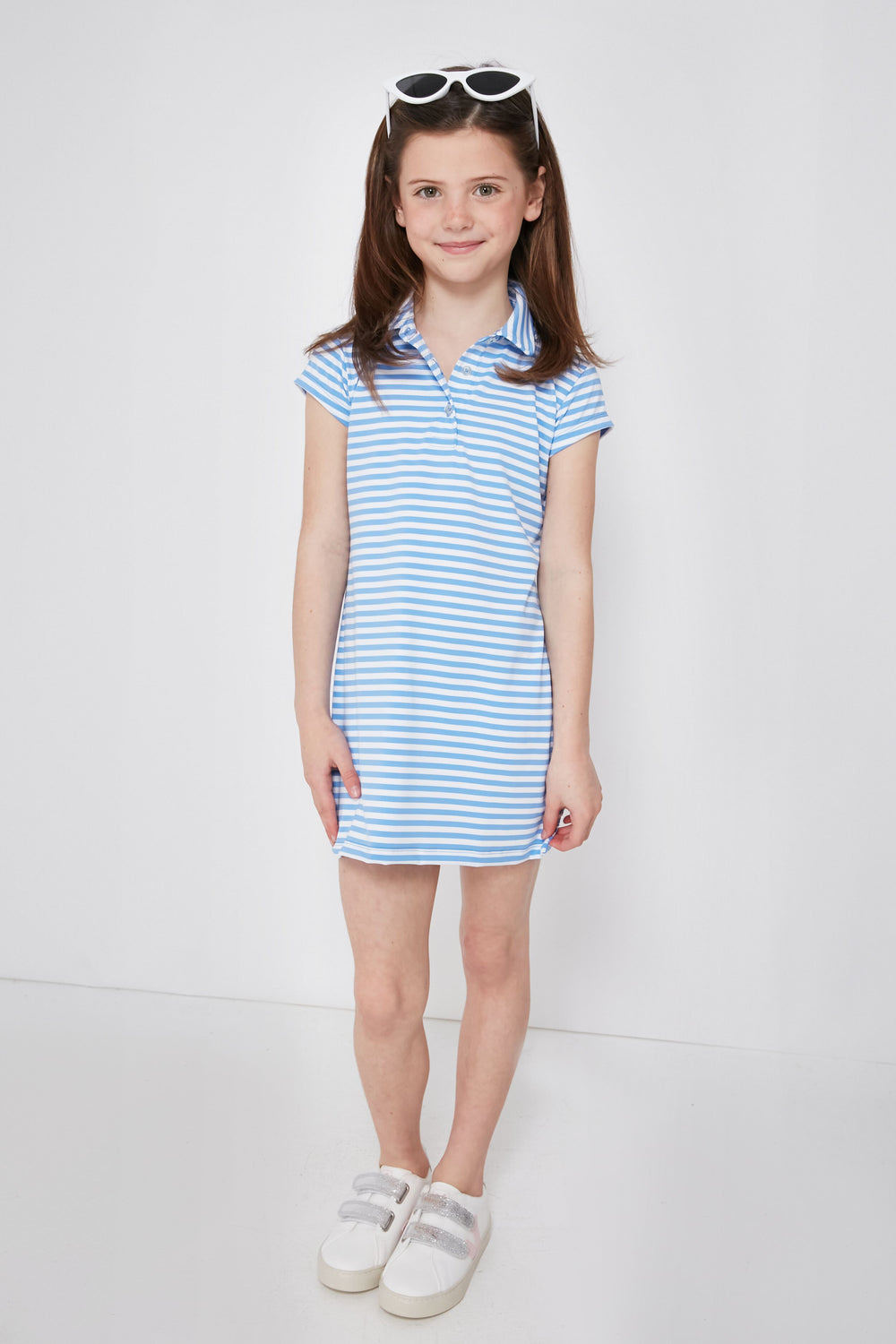 A young girl with brown hair in pigtails wears a Renwick blue and white striped girls' polo dress made from performance fabric, paired with white sneakers and white sunglasses on her head.