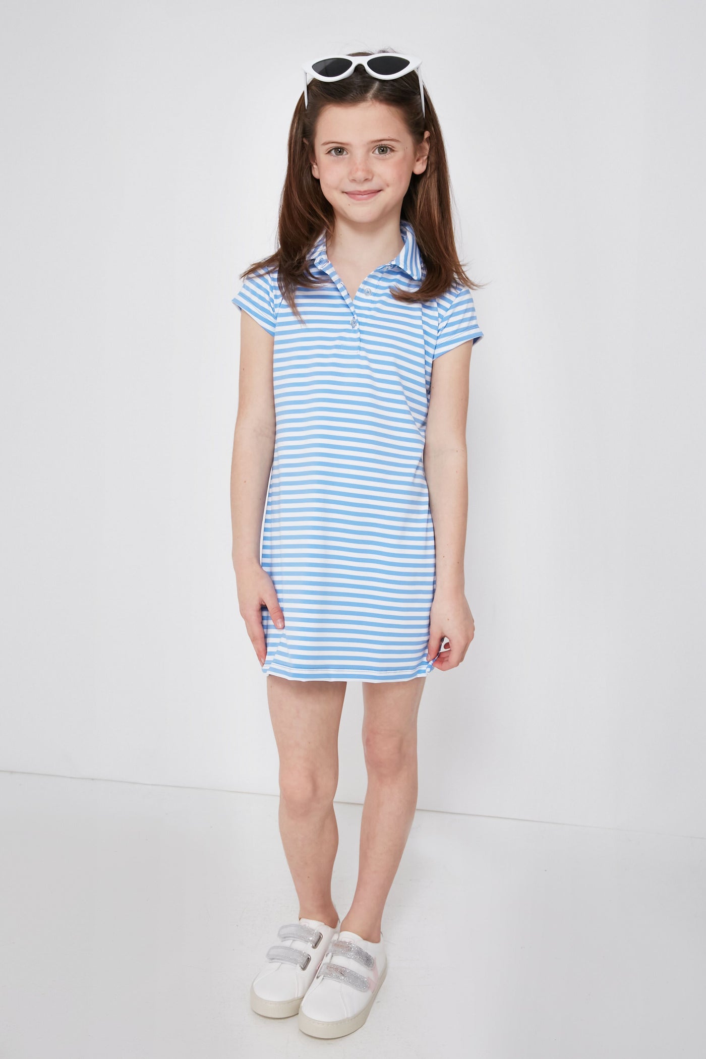 A young girl with brown hair in pigtails wears a Renwick blue and white striped girls' polo dress made from performance fabric, paired with white sneakers and white sunglasses on her head.