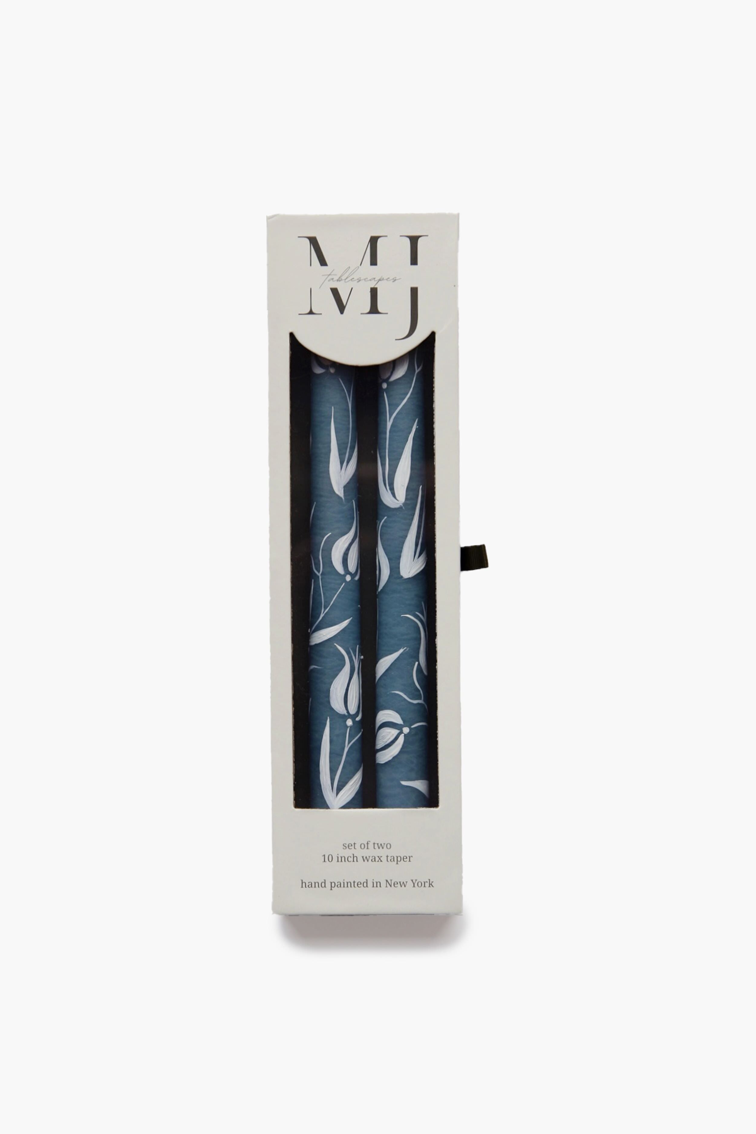 A box of two blue tulip candles with a white floral pattern from MJ Tablescapes, partially visible through a clear window. The packaging features printed text and MJ Tablescapes branding.