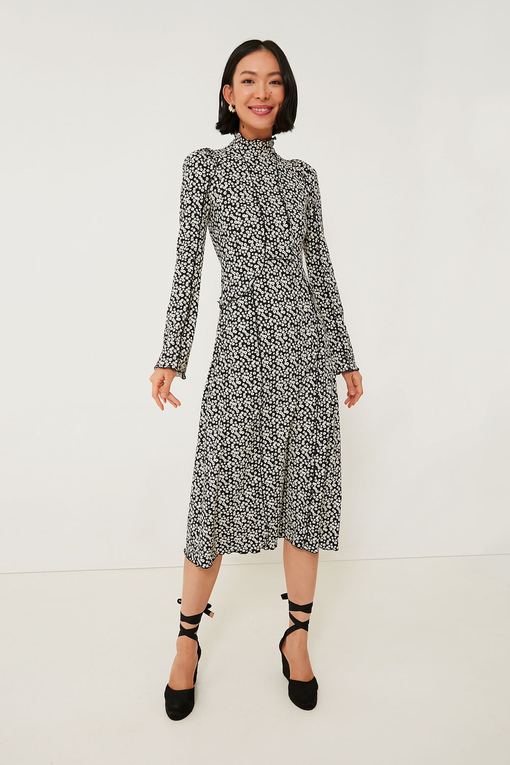 A woman wears a black and white midi ruffle dress with long sleeves by Sea New York, paired with black ankle-tie heels and minimal jewelry.