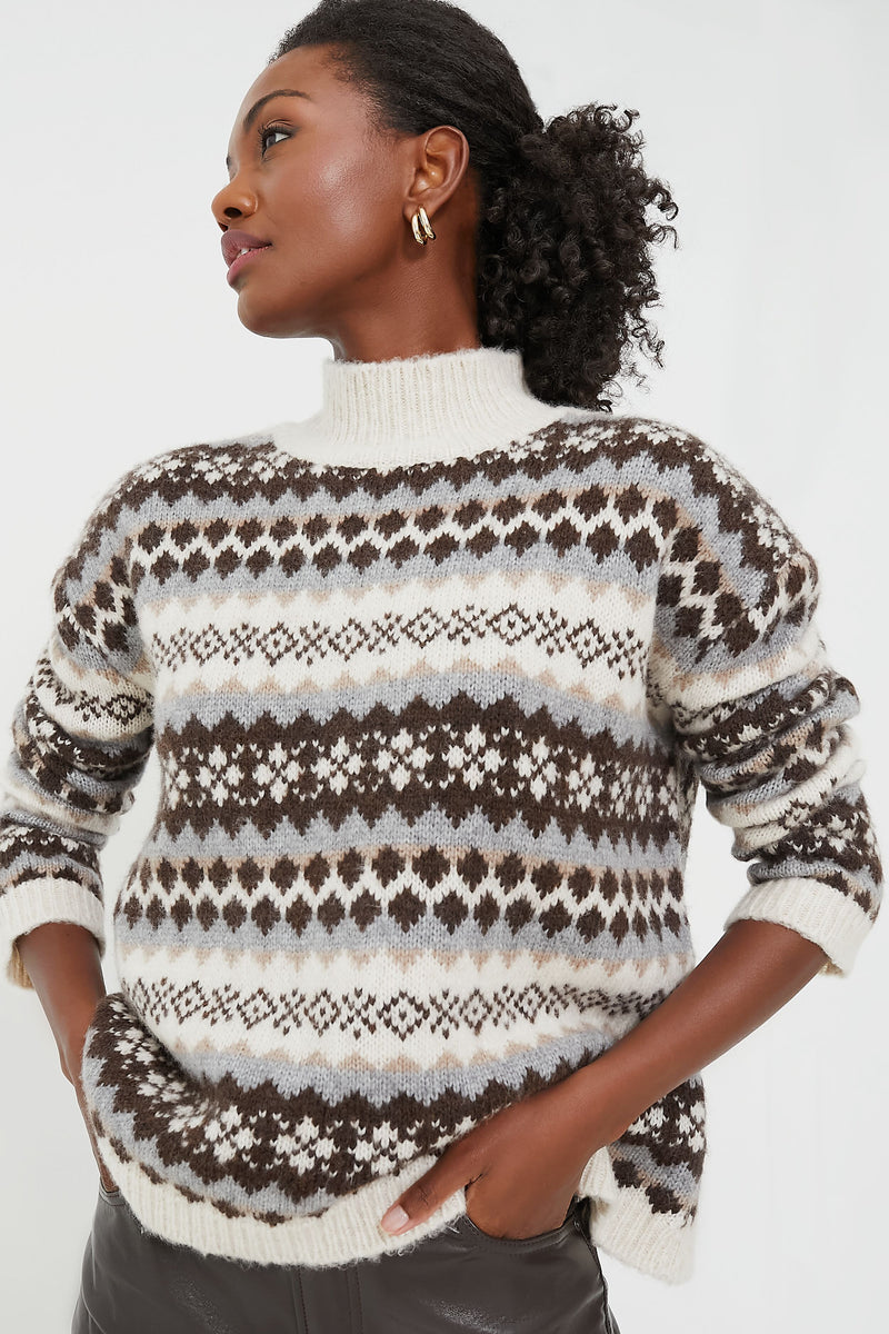 A person wearing a Tuckernuck brown and grey Fair Isle sweater with geometric patterns stands with one hand in their pocket, gazing to the side.