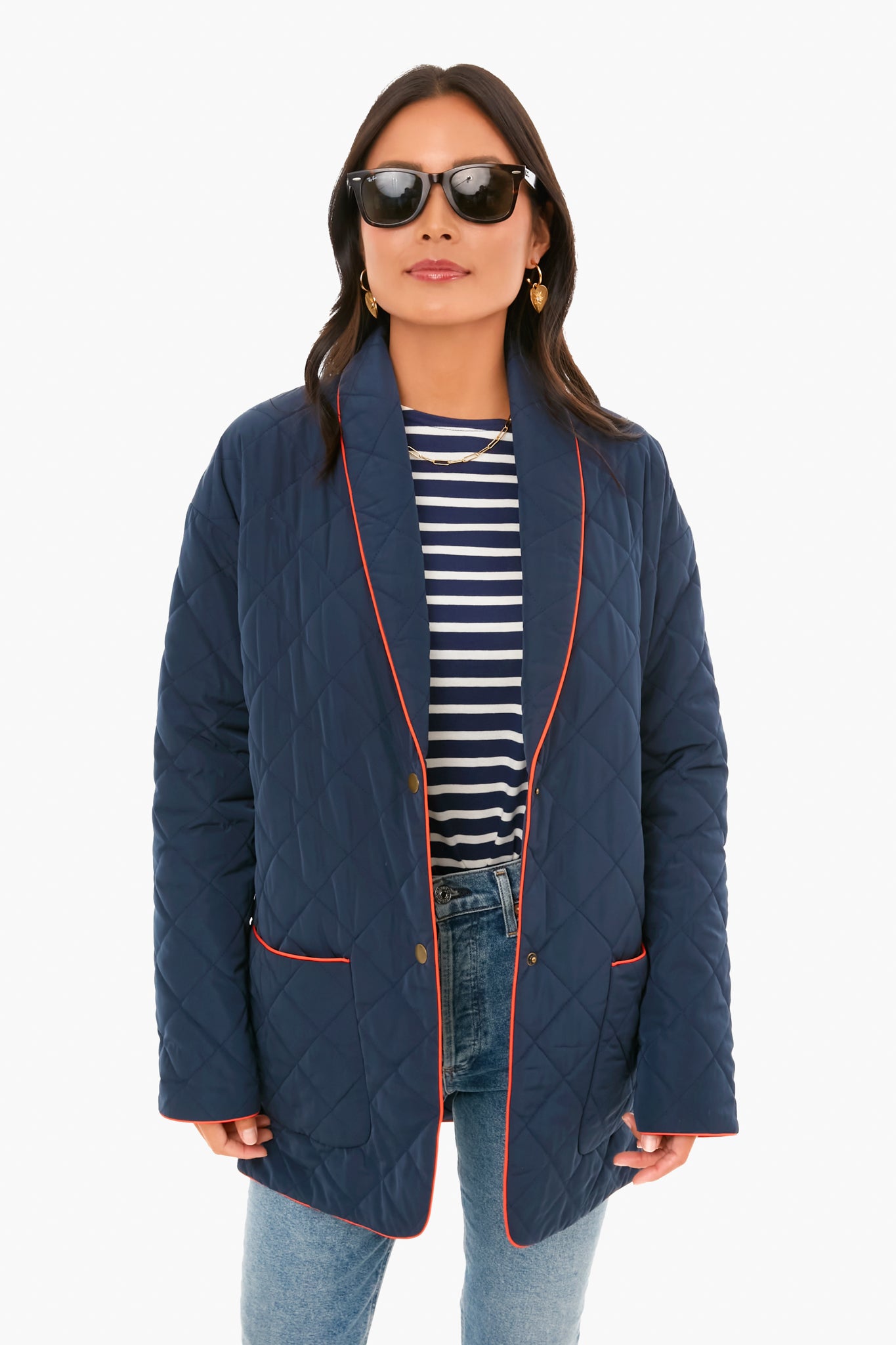 A woman wears KULE sunglasses, a navy quilted jacket with red piping, a striped tee, and blue jeans.