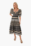 A woman smiles in a Sea New York black and beige bohemian print dress with puff sleeves and a keyhole neckline, styled with heeled sandals and a necklace.