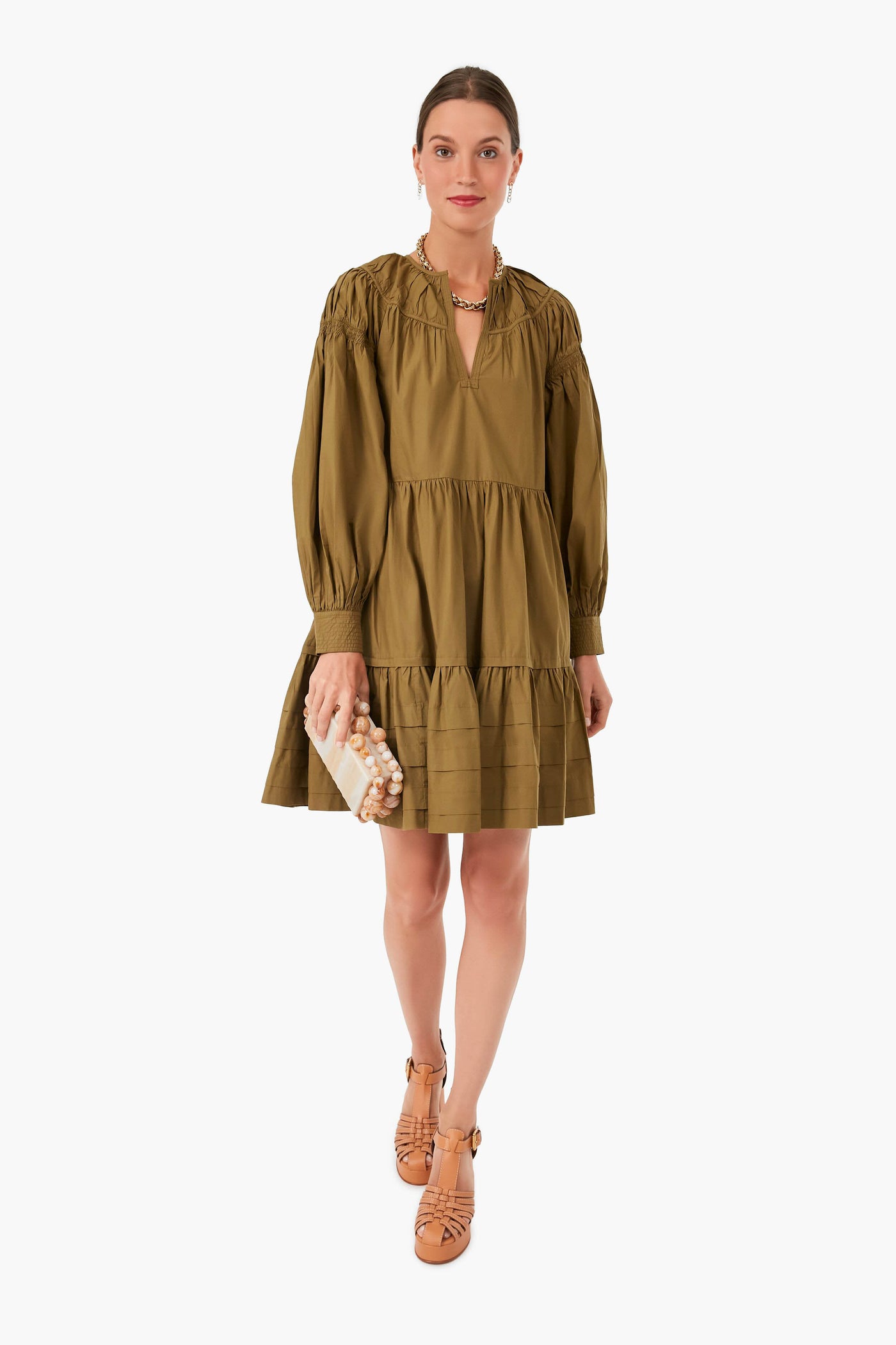 A woman in an olive green Ulla Johnson long-sleeve tiered mini dress, paired with tan lace-up sandals and a beaded clutch.