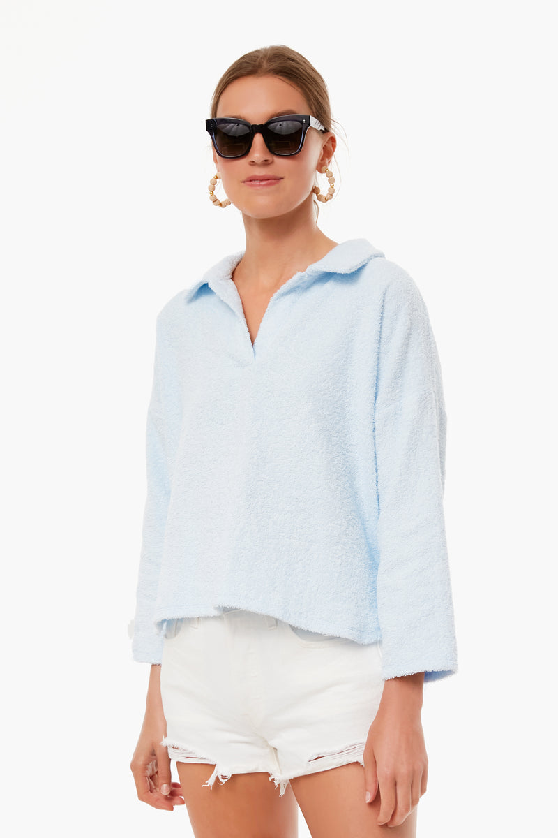 A woman wears a Pomander Place light blue terry cloth polo long-sleeve top with white ripped shorts, large hoop earrings, and black sunglasses—ideal for chic beachwear or as a stylish coverup.