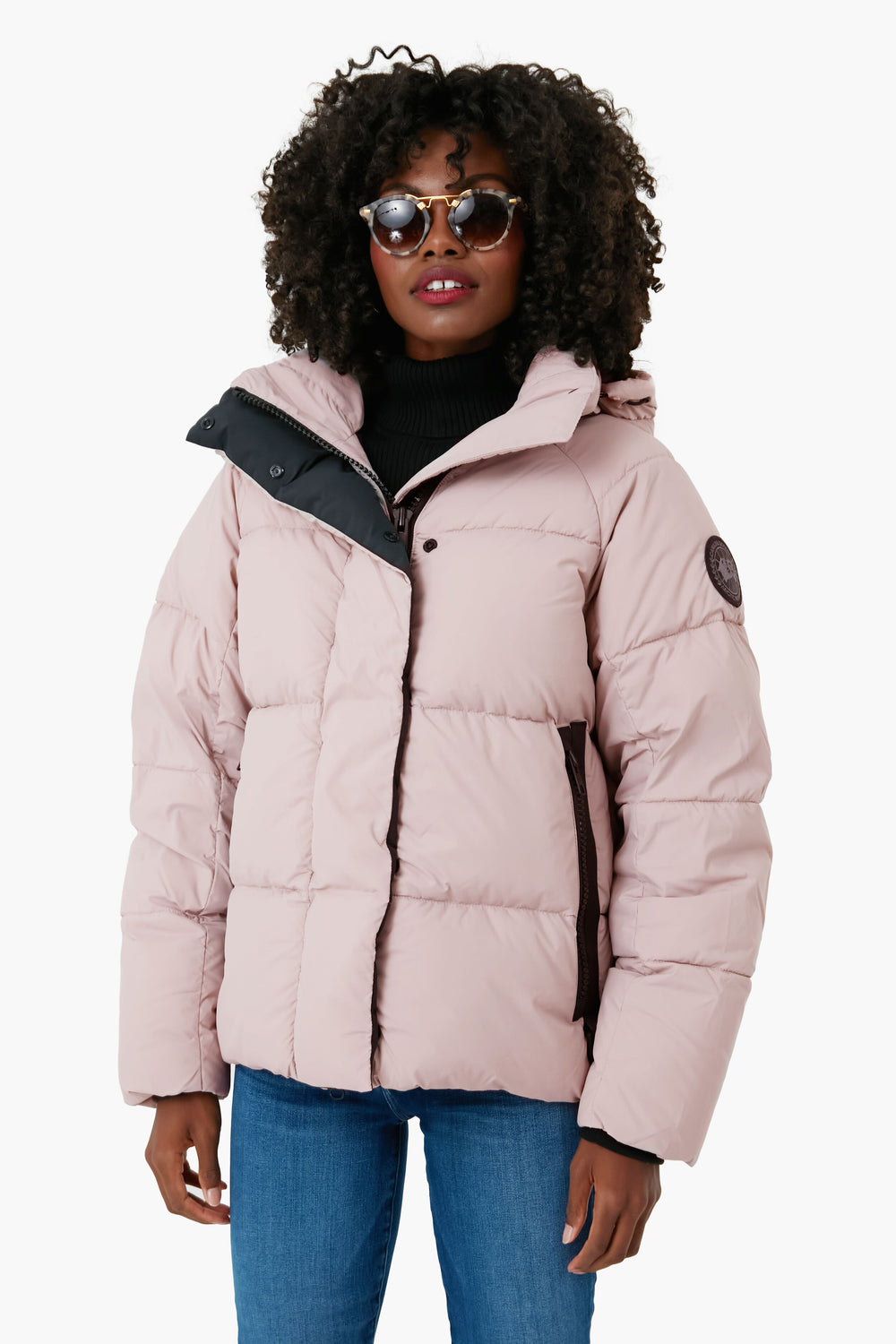 A person stands confidently in sunglasses, a black turtleneck, blue jeans, and a light pink hooded Canada Goose jacket.
