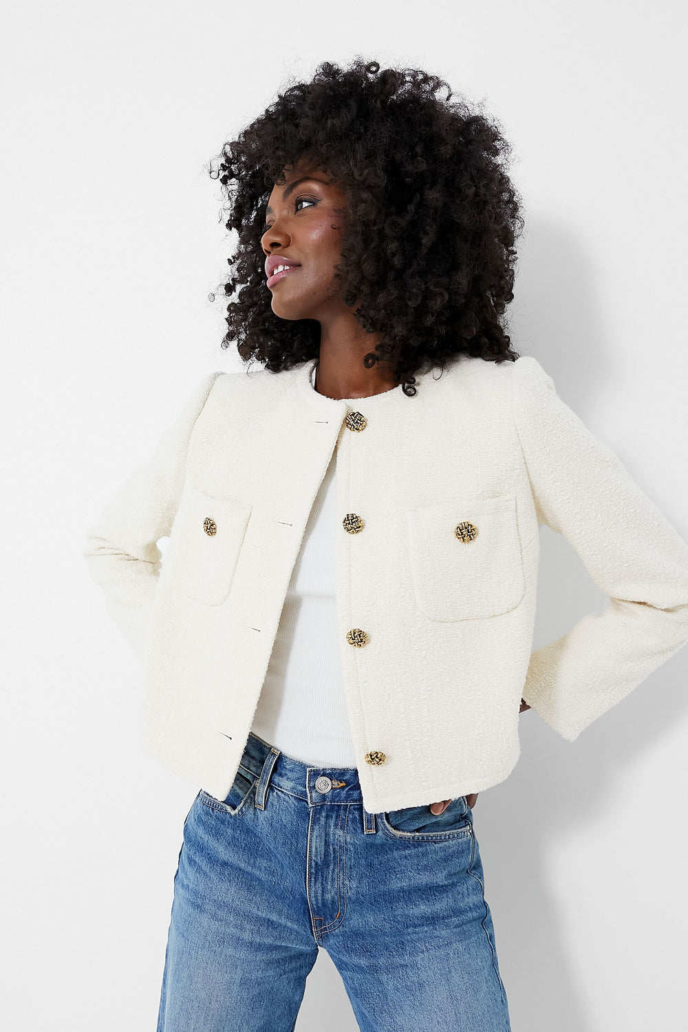 A woman wears a ba&sh Meredith Jacket—an ecru textured tweed cropped jacket with large gold buttons—over a white top and blue jeans, evoking a chic French vibe.
