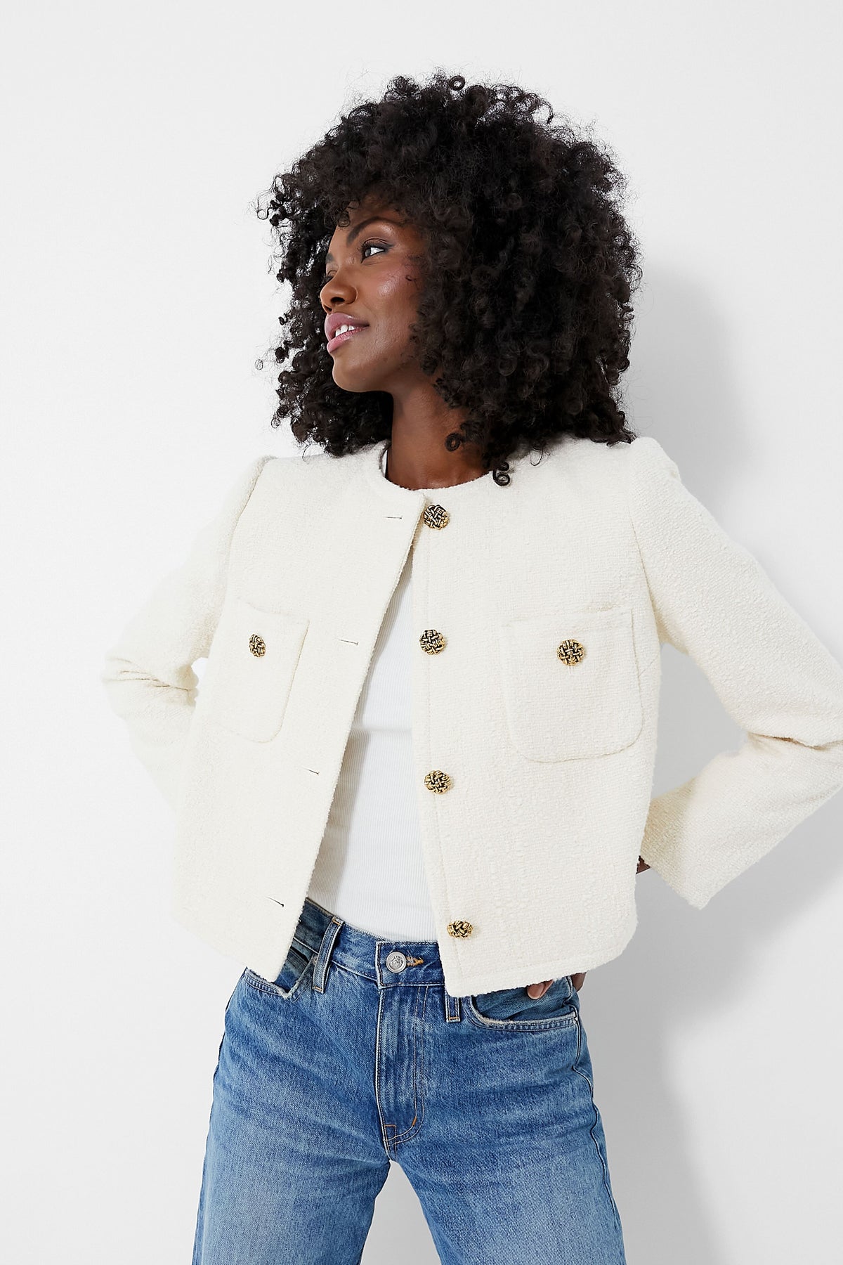 A woman wears a ba&sh Meredith Jacket—an ecru textured tweed cropped jacket with large gold buttons—over a white top and blue jeans, evoking a chic French vibe.