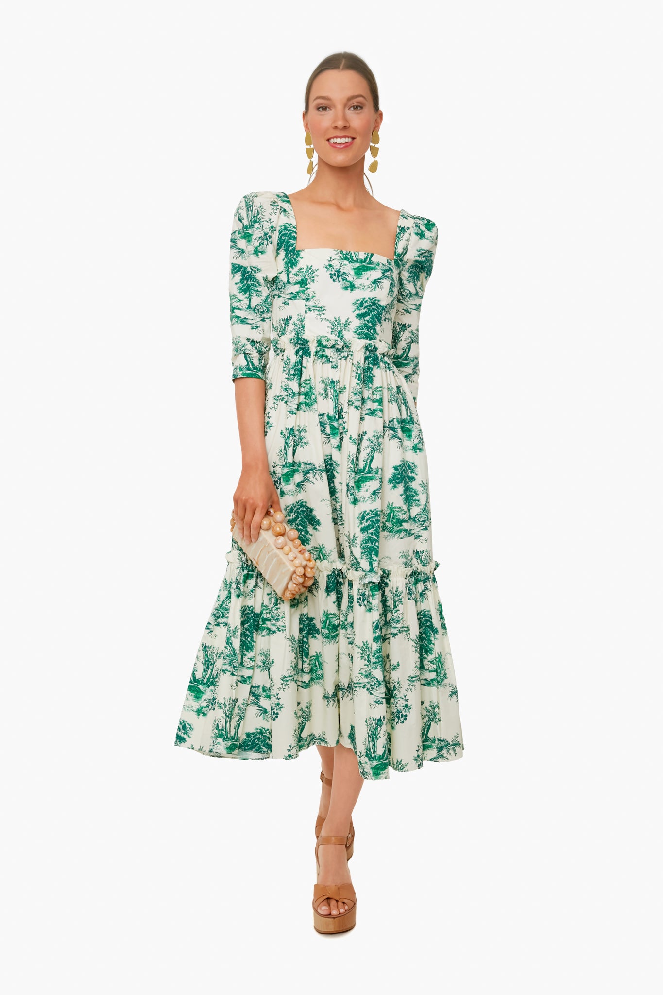 A woman wears a Cara Cara botanical print A-line midi dress with puffed sleeves, styled with a beige clutch, tan platform sandals, and gold earrings.