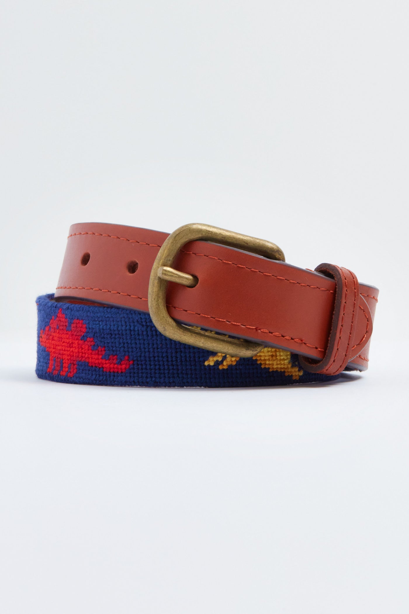 A brown leather belt with a brass buckle and navy needlepoint showing red and yellow dinosaurs, this Smathers and Branson dinosaur belt is perfect for children.