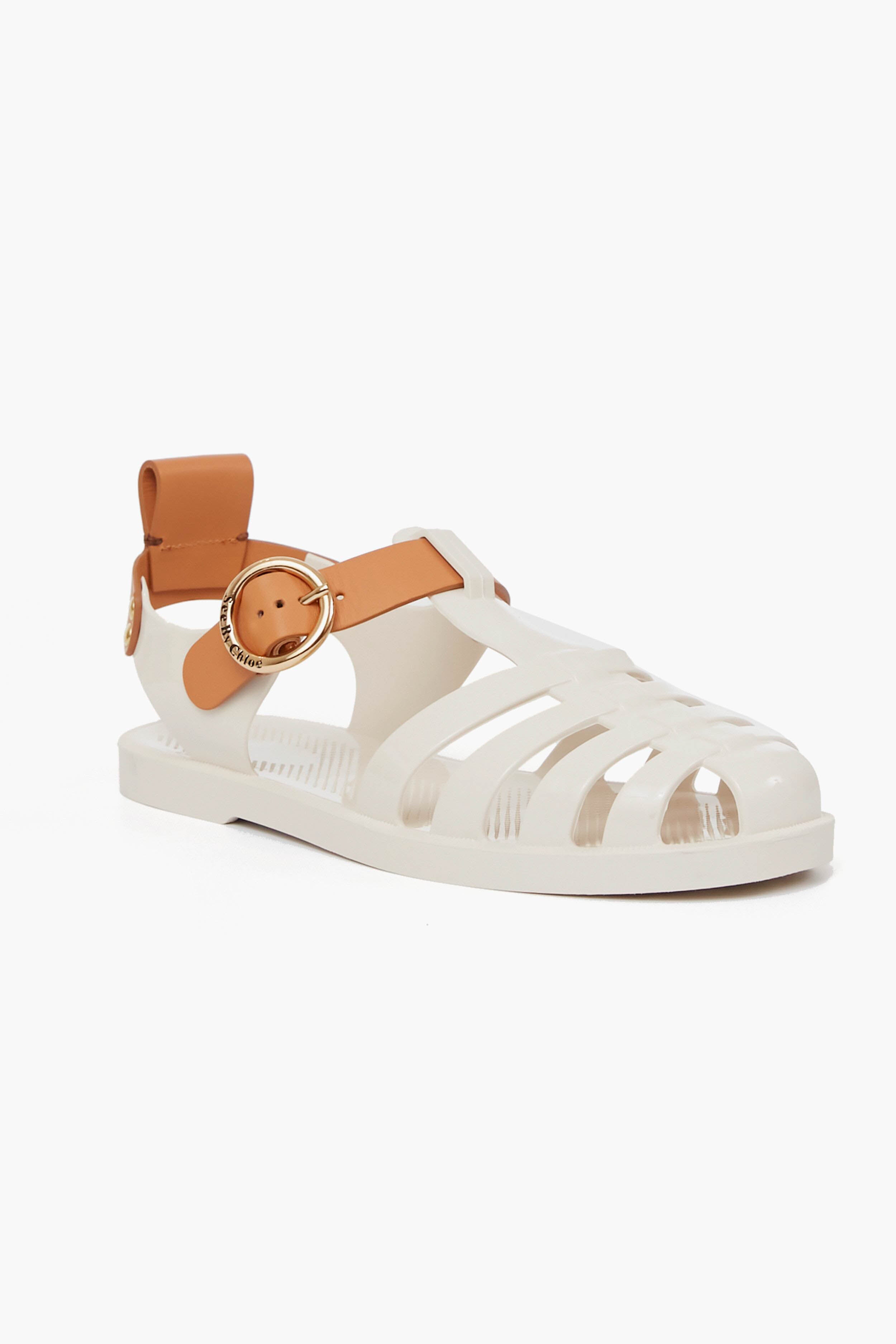 See by Chloe presents a white jelly fisherman sandal featuring a tan ankle strap, gold buckle, and cut-out design on the upper and sides, inspired by '70s footwear.