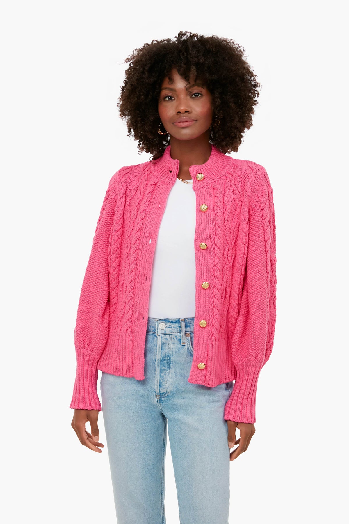A woman wears a bright pink Tuckernuck chunky cardigan over a white top and light blue jeans, combining cozy comfort with stylish flair.