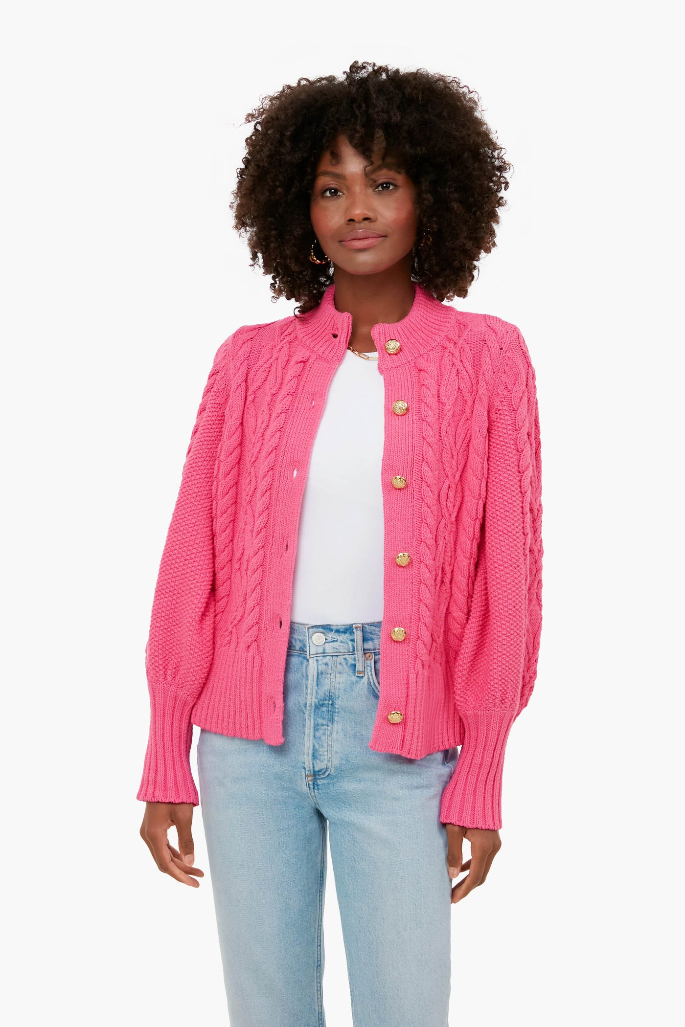 A woman wears a bright pink Tuckernuck chunky cardigan over a white top and light blue jeans, combining cozy comfort with stylish flair.