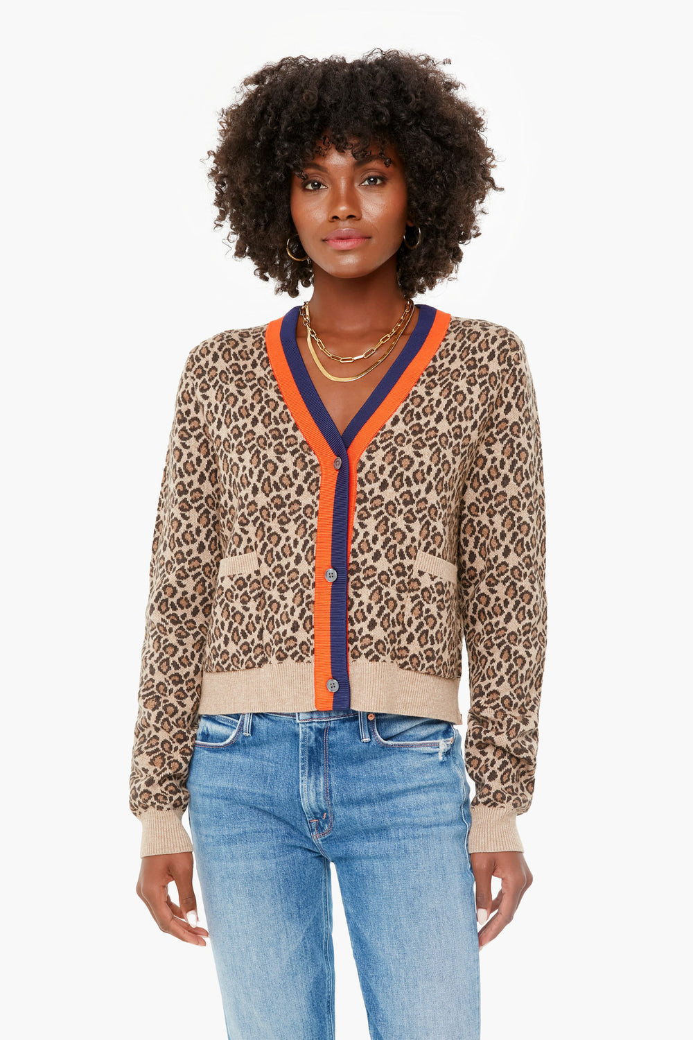 A woman effortlessly showcases the KULE Leopard Milo Cardigan with orange and navy trim, paired with blue jeans and layered gold necklaces, making it a standout statement piece.