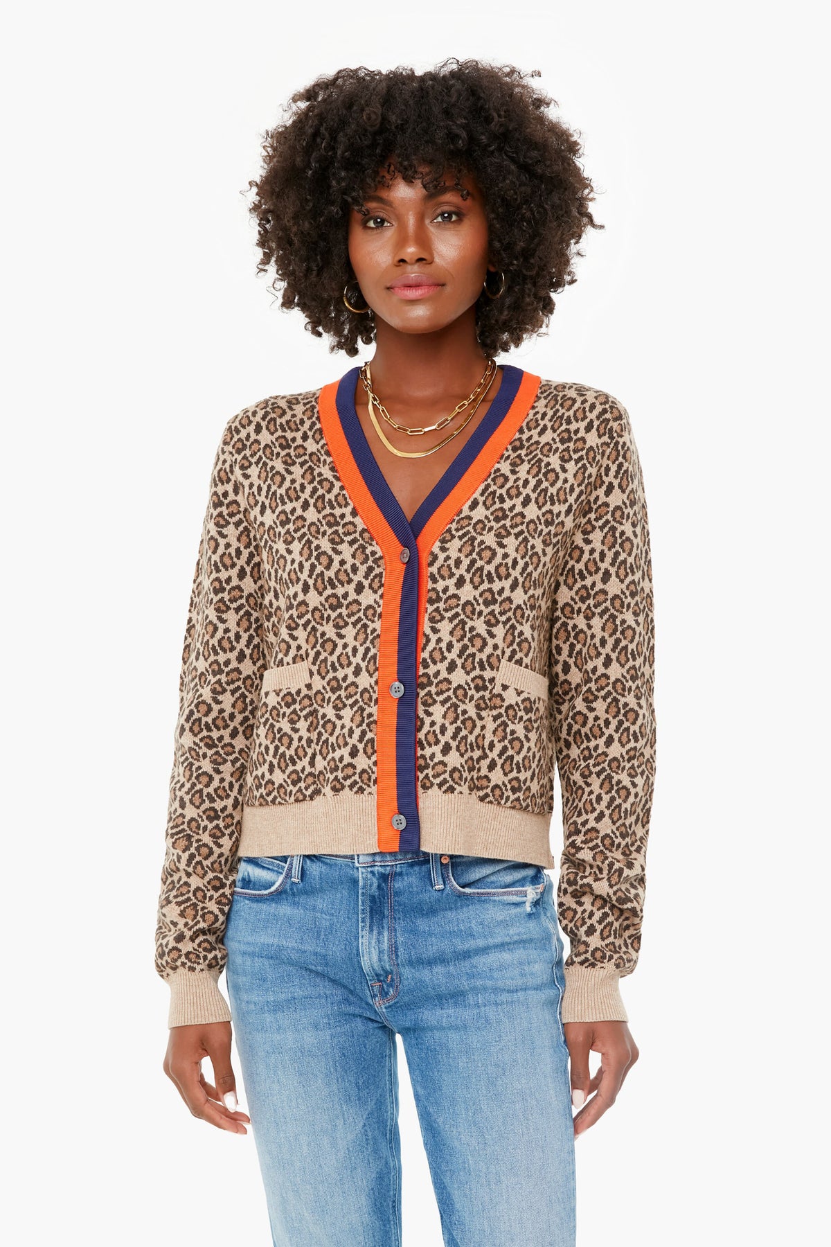 A woman effortlessly showcases the KULE Leopard Milo Cardigan with orange and navy trim, paired with blue jeans and layered gold necklaces, making it a standout statement piece.