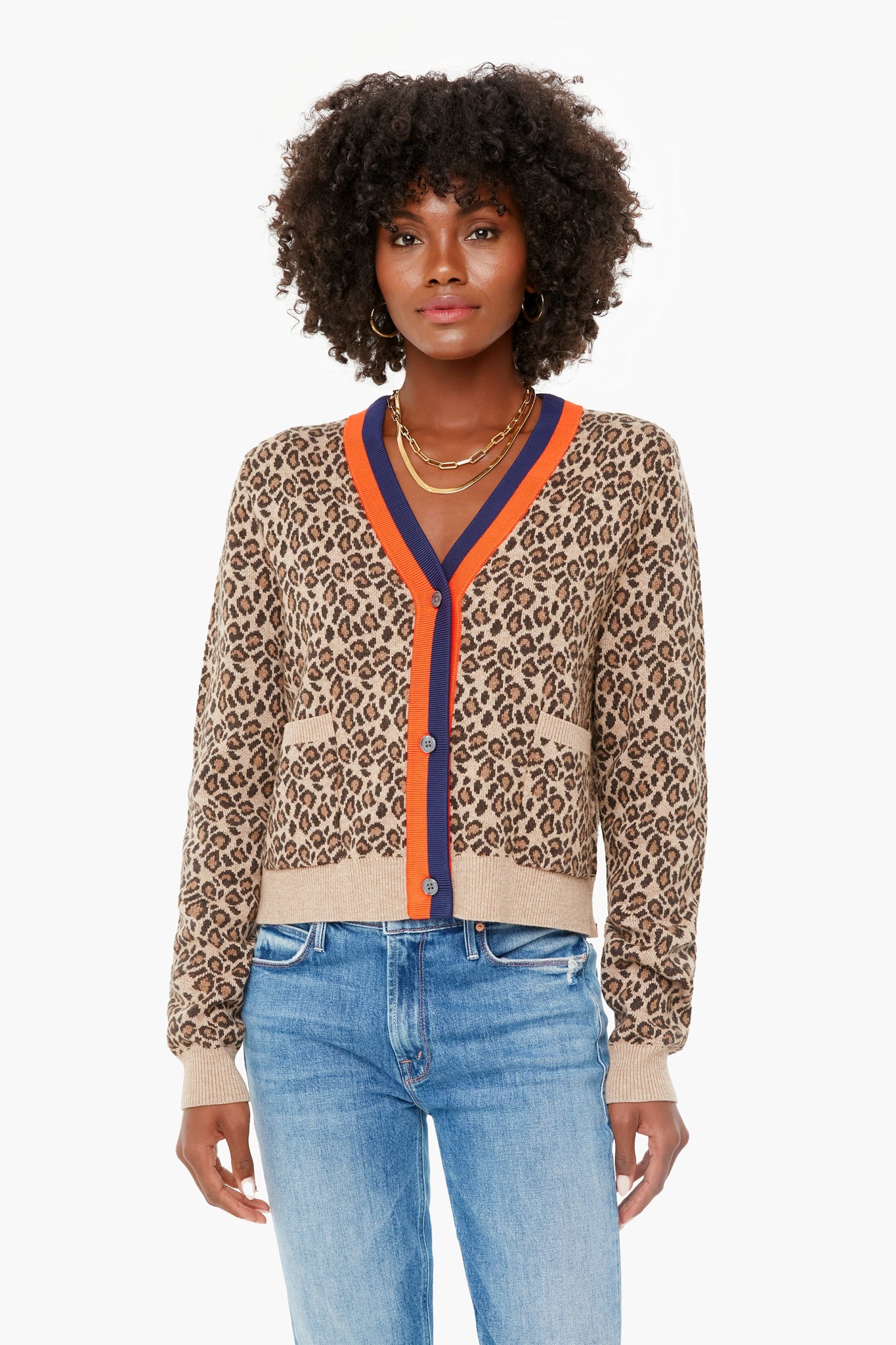 A woman effortlessly showcases the KULE Leopard Milo Cardigan with orange and navy trim, paired with blue jeans and layered gold necklaces, making it a standout statement piece.