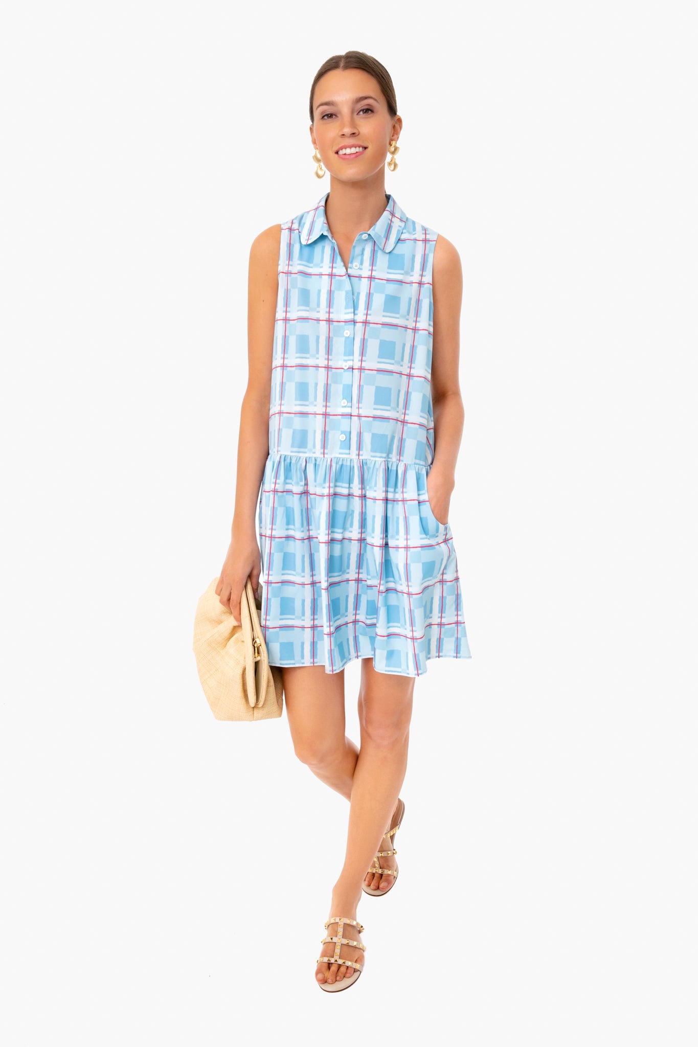 A woman is smiling in a Tuckernuck Sleeveless Hayes Shirt Dress in blue plaid with a collared neckline and drop waist, accessorized with a beige clutch and sandals.